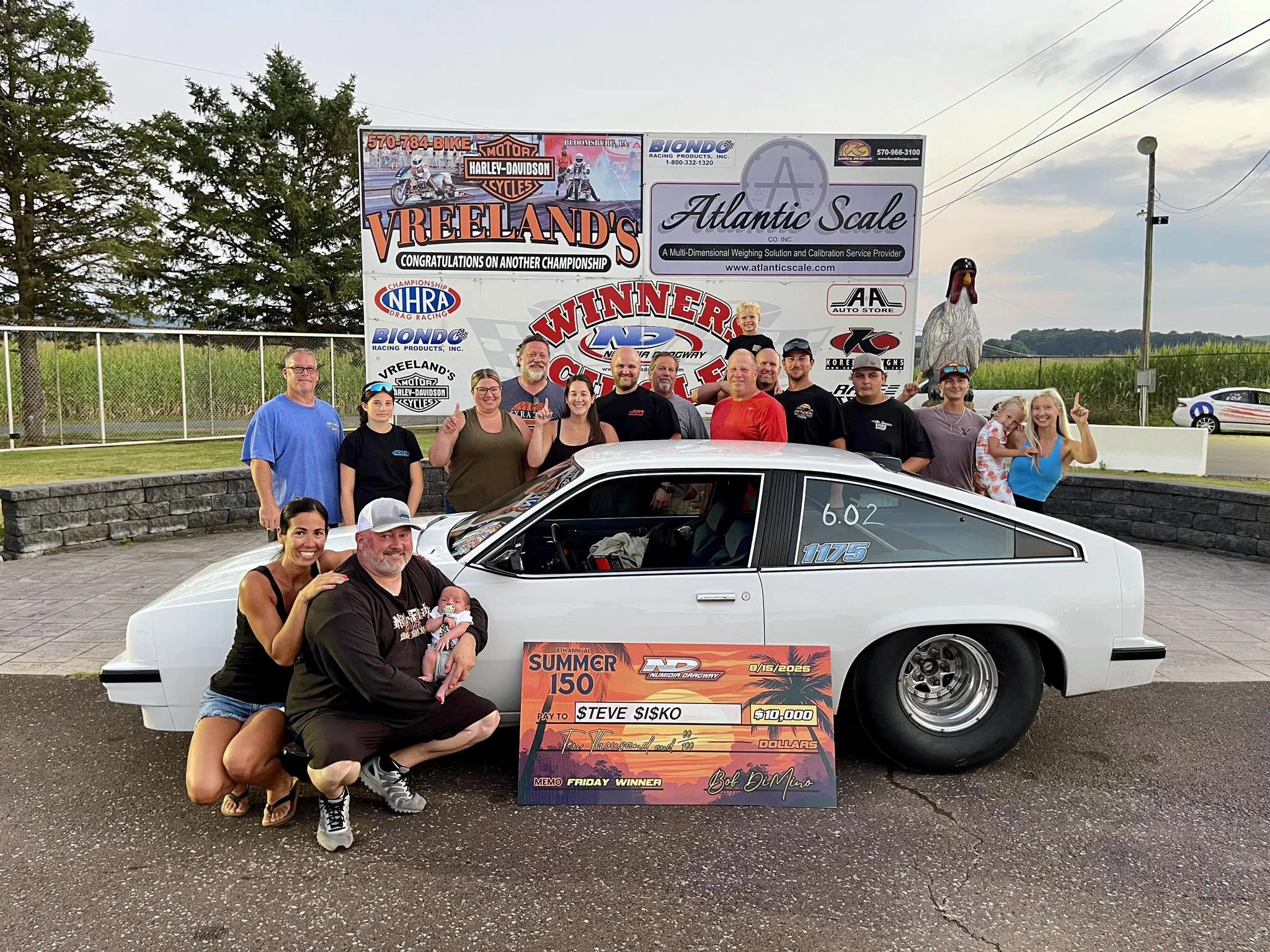 A group of people celebrating next to a white race car with a large check for $10,000 in front of a signboard at a racing event. The signboard displays various sponsors and indicates a victory at the Summer 150 race at Nomad Raceway on August 15, 202