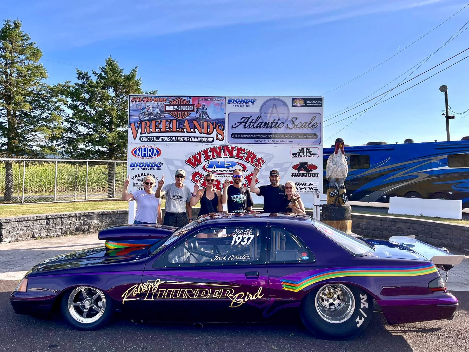 A group of five people celebrating at a car racing event, standing behind a black race car with rainbow-colored stripes and the name 'Thunder Bird' on the side, at a track with sponsor banners and a chain-link fence in the background.