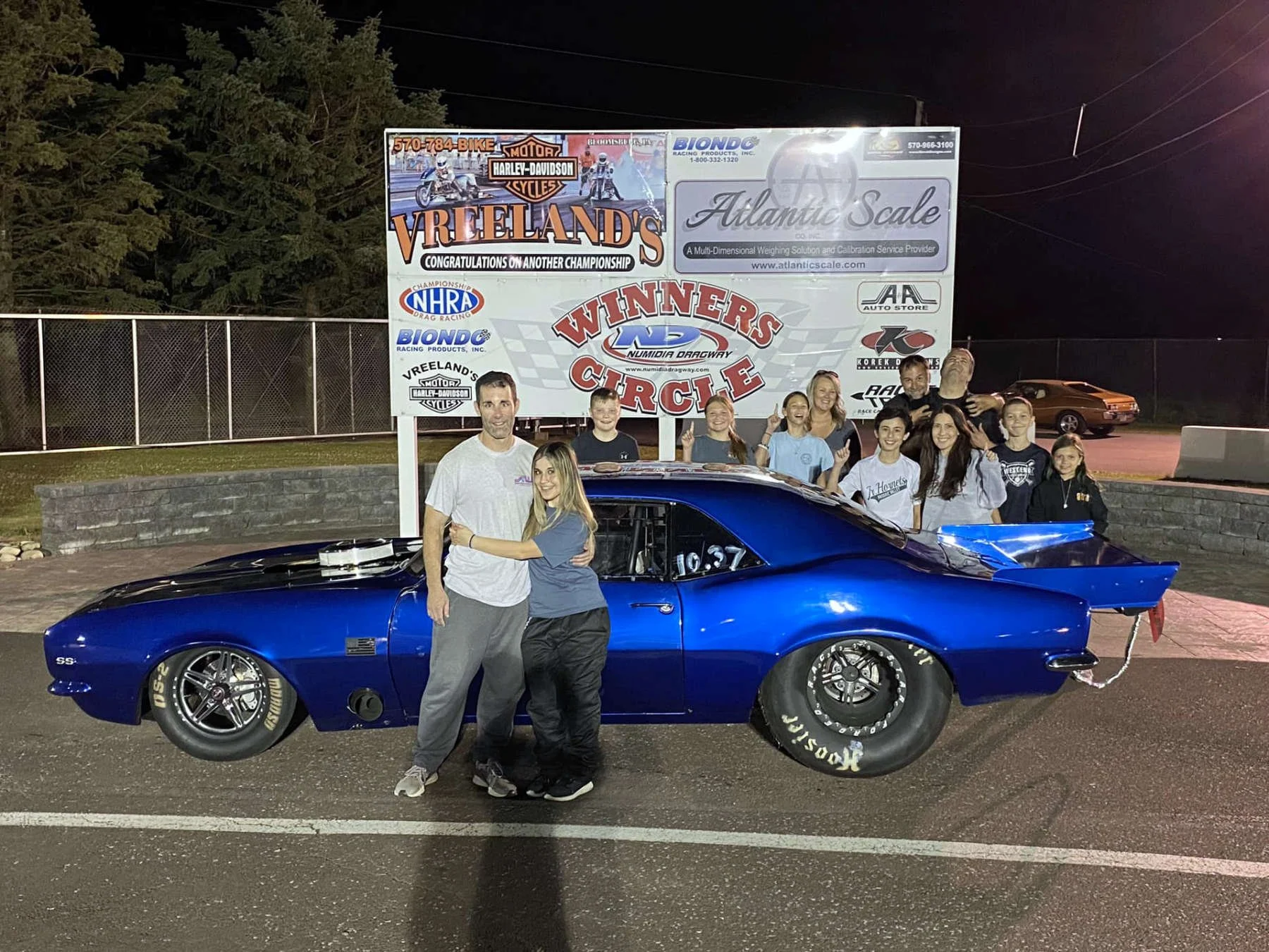 A group of people celebrating near a blue race car at night, with a large sign promoting motorcycle and drag racing championships in the background.