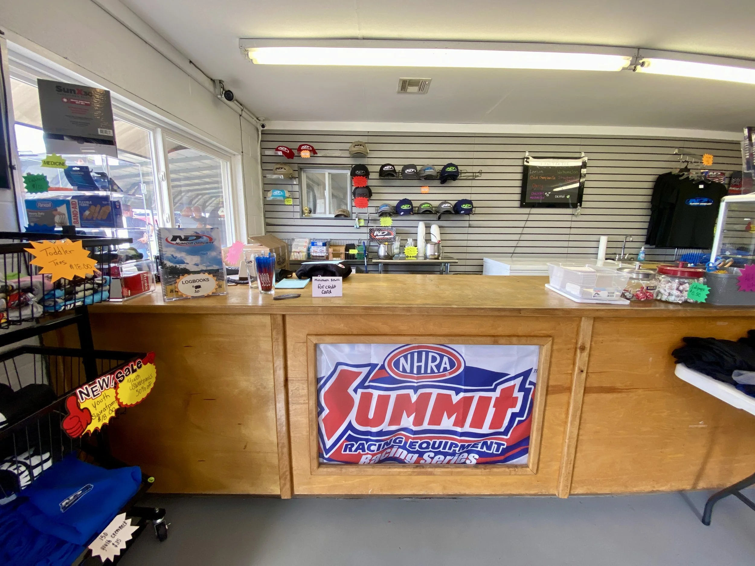 Inside a retail store with helmets and racing equipment, promotional signs on a wooden counter, and NHRA Summit Racing banner front and center.