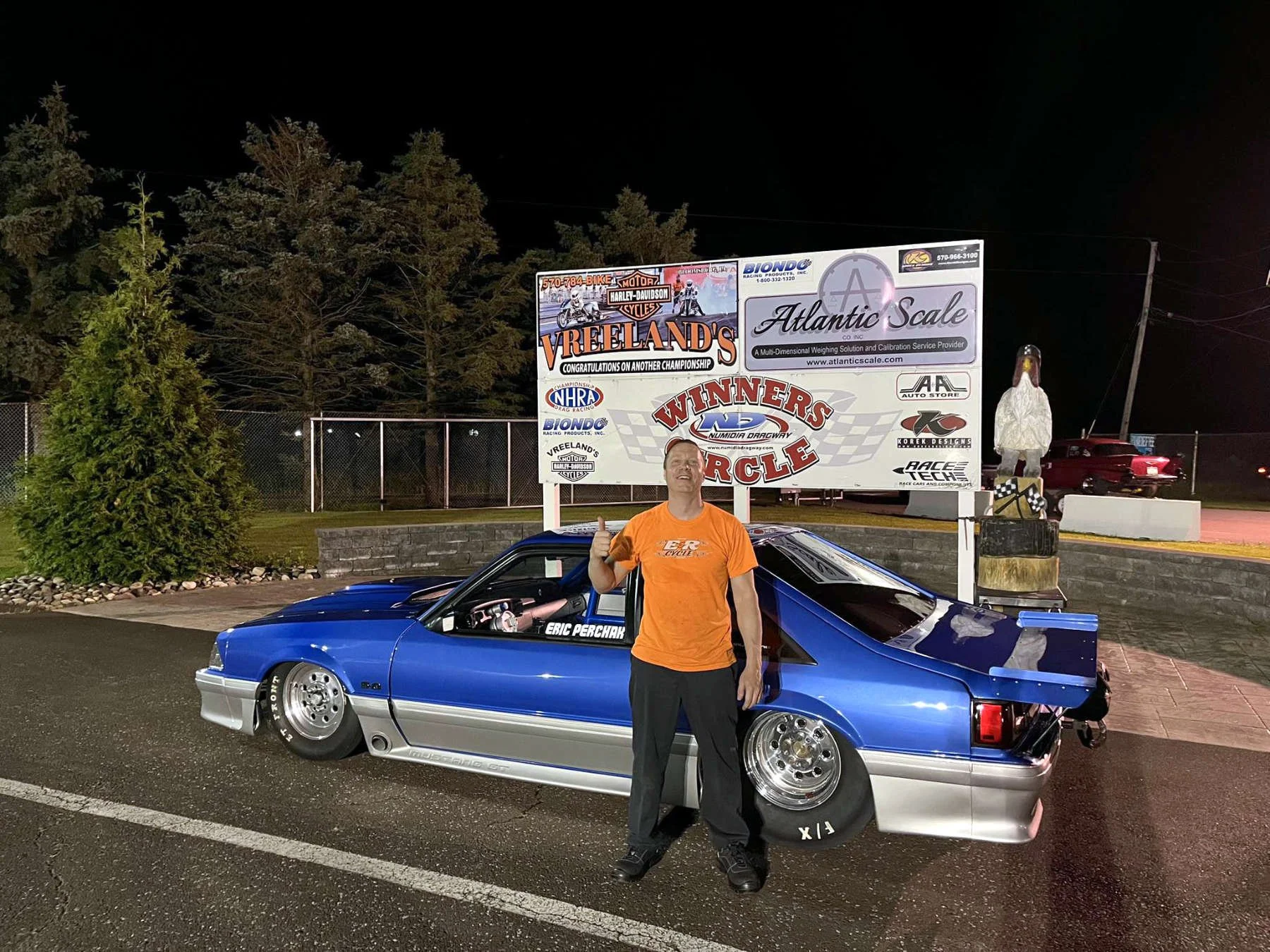 A man in an orange shirt standing next to a blue racing car with custom wheels at a race track. The man is smiling and giving a thumbs up. There are signs in the background with logos and text, including 