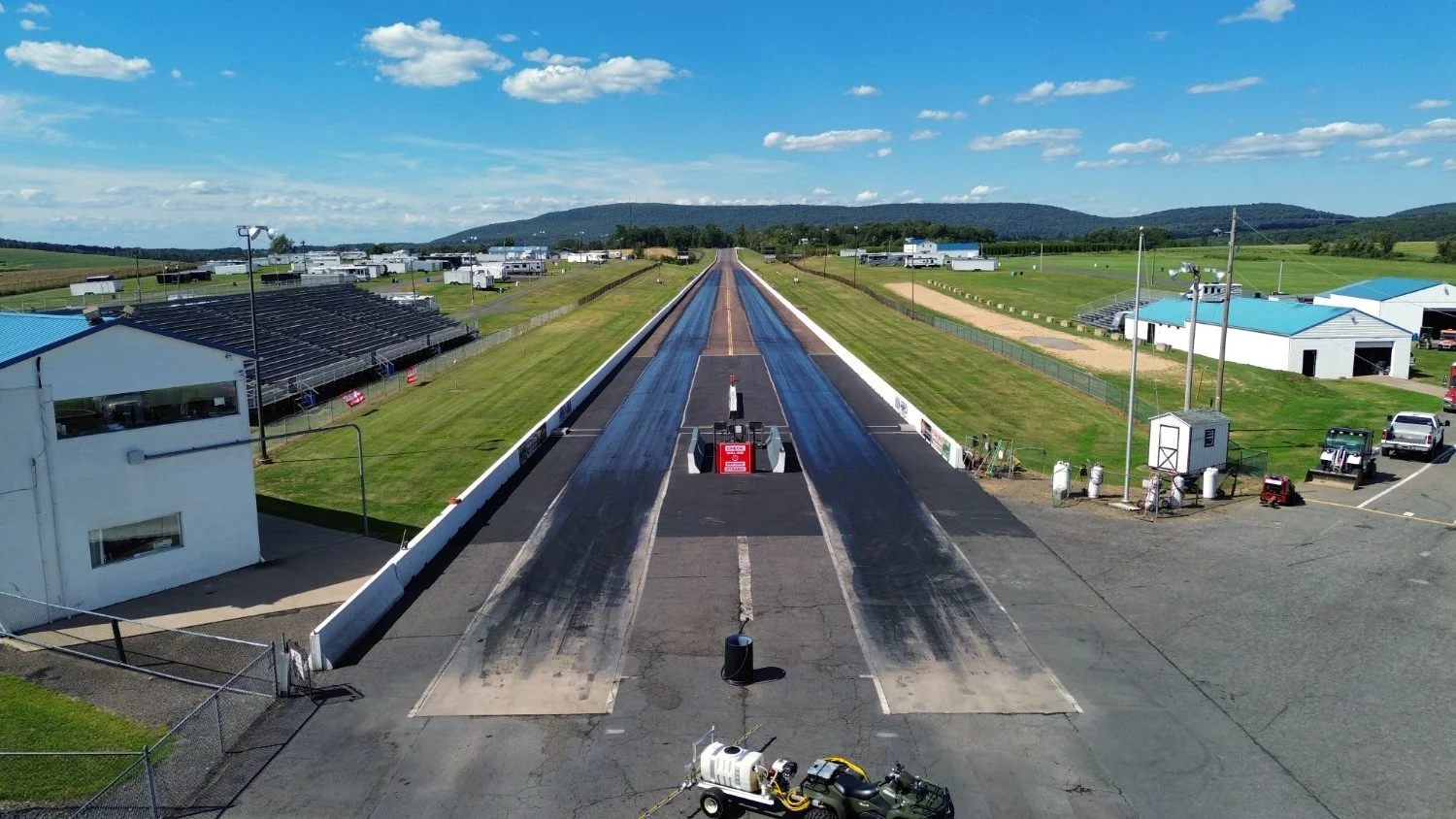 A drag racing strip with a car at the starting line and some equipment nearby, surrounded by a rural area with small buildings and mountains in the background.