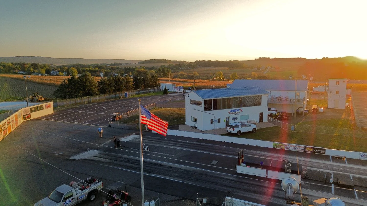 A race track with a starting line, a large American flag on a flagpole, and a building with a glass front at sunset, surrounded by a parking lot and rural landscape.