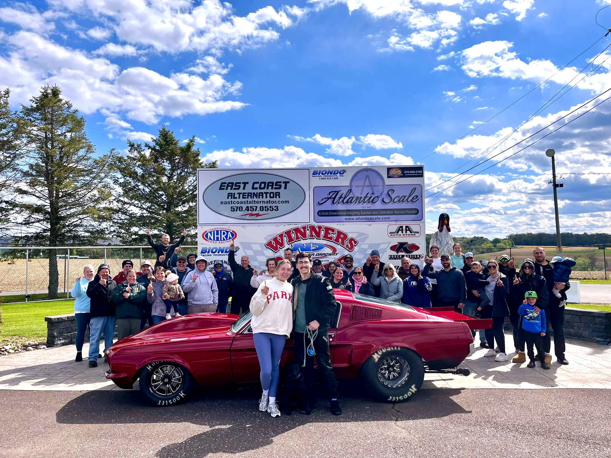 A large group of people gathered outdoors, some standing behind a red vintage car, posing for a photo with a bright, partly cloudy sky overhead. A winner circle with racing-related logos and sponsors is visible in the background.