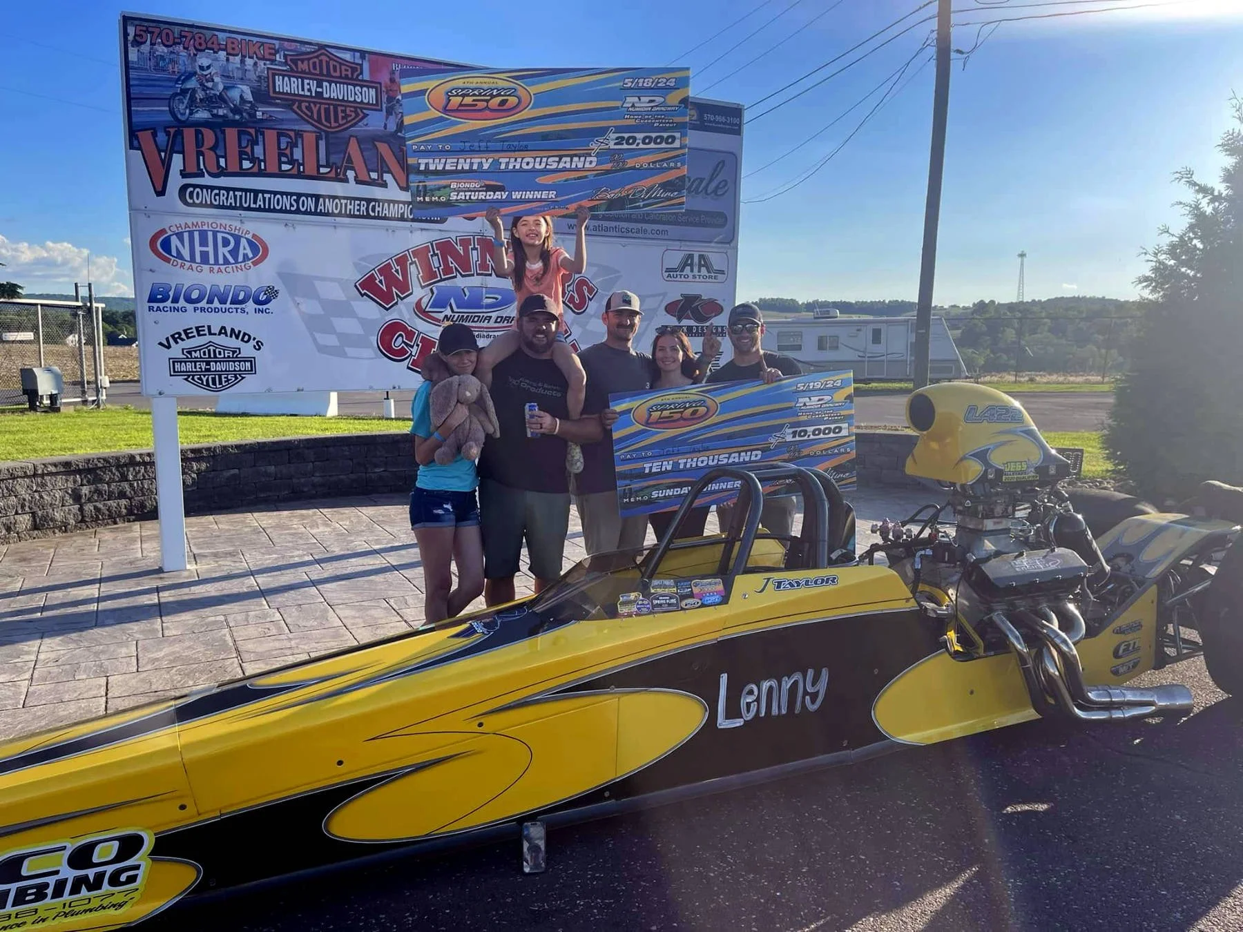 A group of people celebrating victory at a drag racing event. A young girl is sitting on a man's shoulders, and they are holding a large check for ten thousand dollars. A yellow drag racing vehicle with the name 'Lenny' on it is in the foreground. Th