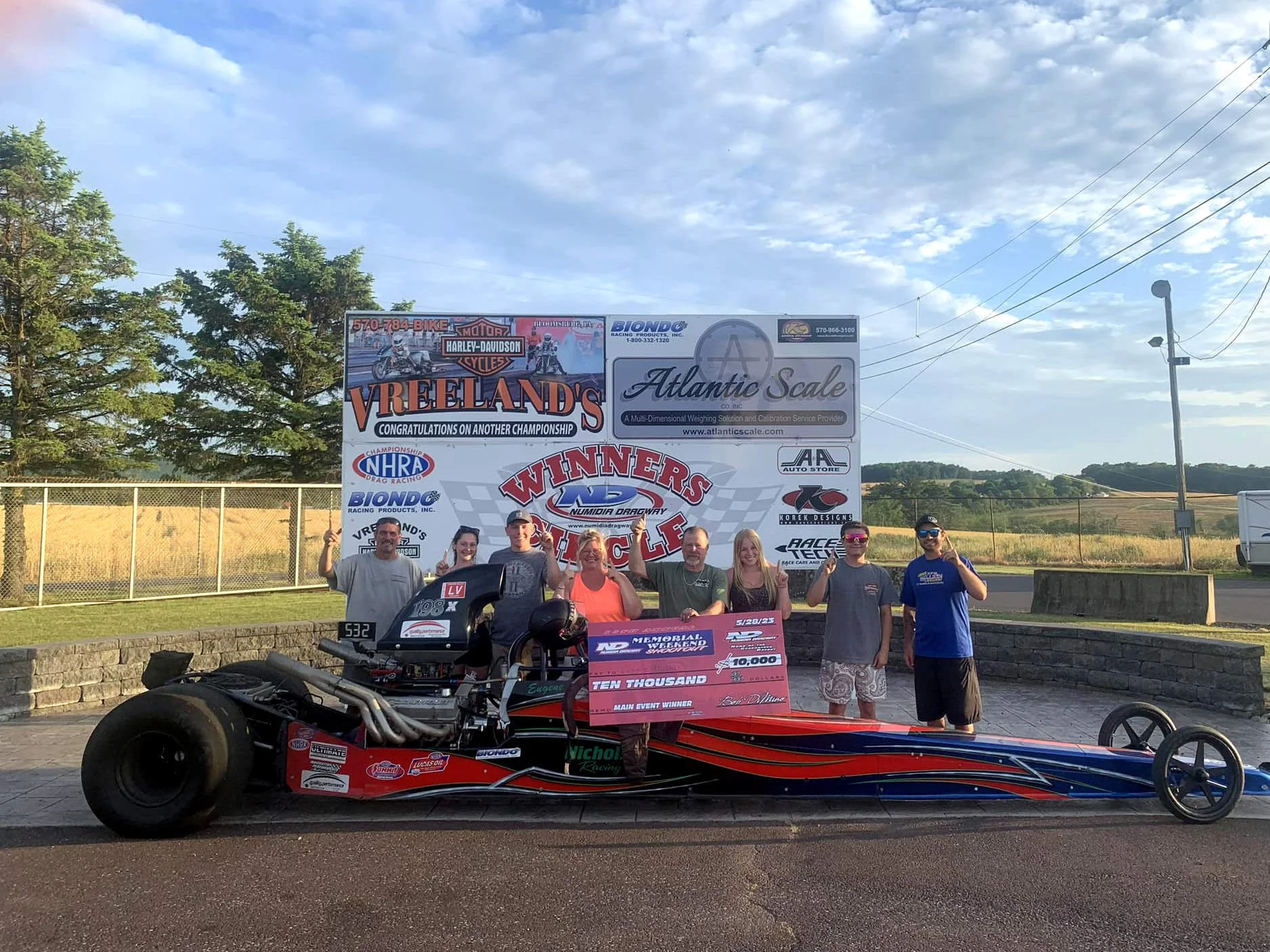 A group of people standing behind a racing car at an outdoor event, with a large banner in the background that reads "Vreeland's" and "Atlantic Scale." The setting appears to be a race track or event venue with a fence, trees, and hills under a partl