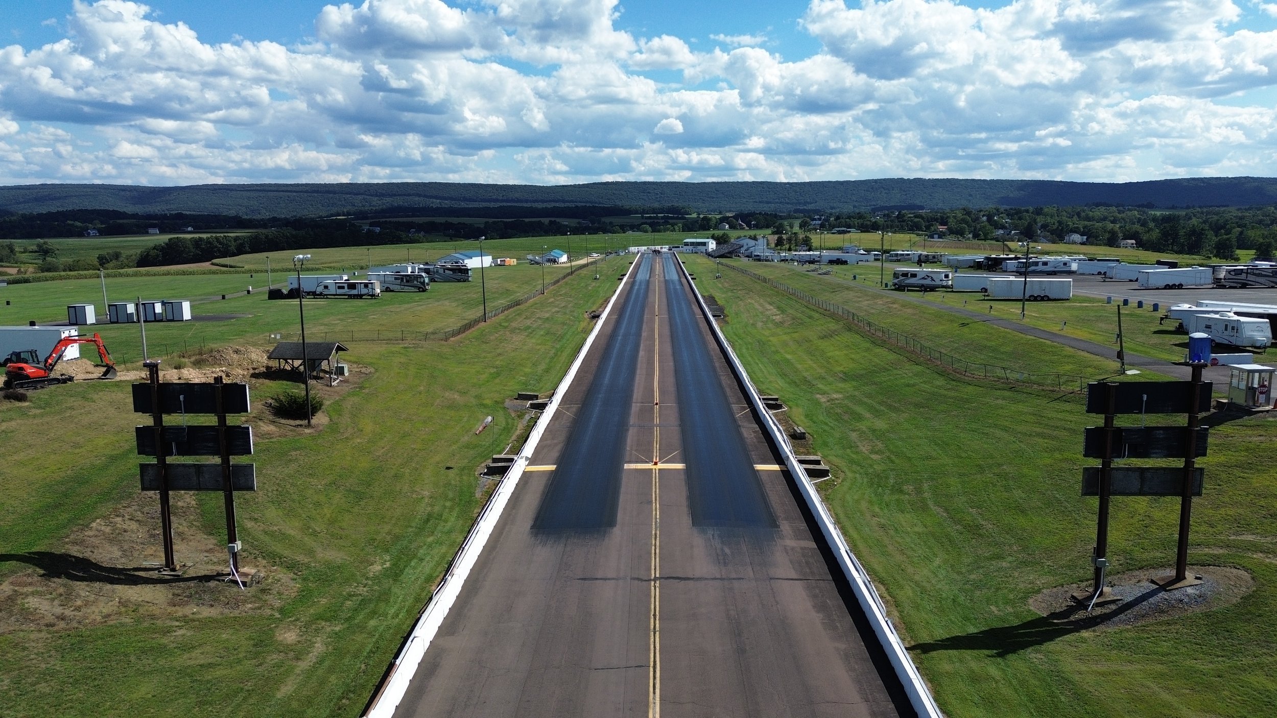 View of a drag strip with a long starting line under construction, surrounded by grassy fields, trailers, and recreational vehicles, with a scenic backdrop of hills and a partly cloudy sky.