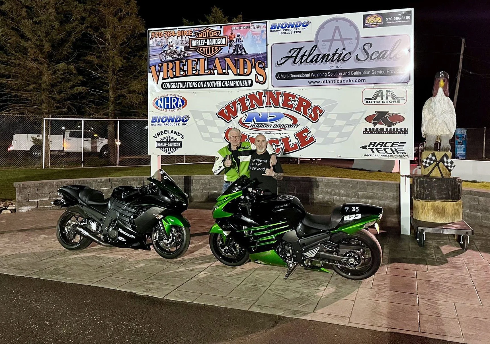 Two men standing behind two Kawasaki motorcycles, one green and one black, in front of a large race track sign that says 'Vreeland's Winners Circle' at night. A statue of a duck on a box stands to the right of the sign and a chain-link fence with vehicles behind it is in the background.
