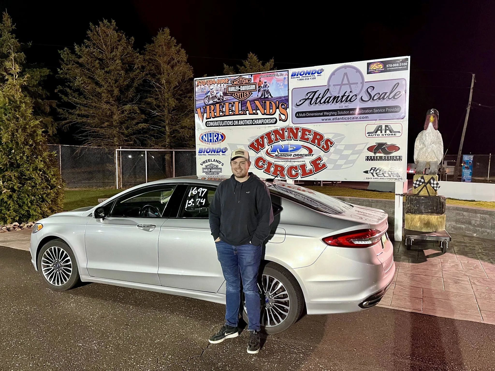 A man standing next to a silver car at night with a large billboard advertising motorcycle and drag racing in the background.