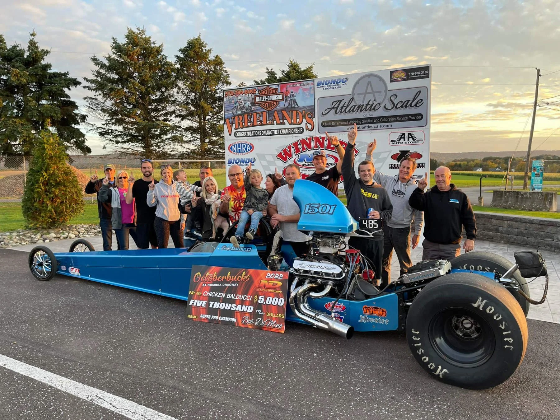 Group of people celebrating around a drag racing car at a victory event, with banners and a sign indicating a $5,000 prize for the Octoberbucks drag race at Numidia Dragway.