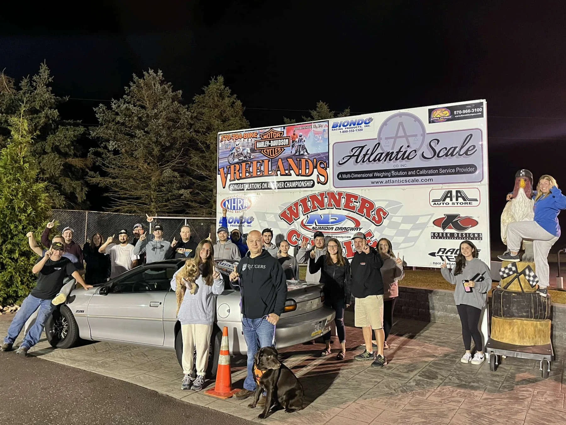 Group of people and dogs standing in front of a racing track billboard at night. The billboard displays advertisements for Atlantic Scale, Harley-Davidson, and other sponsors. The group includes men, women, and children, some holding dogs, with a sil