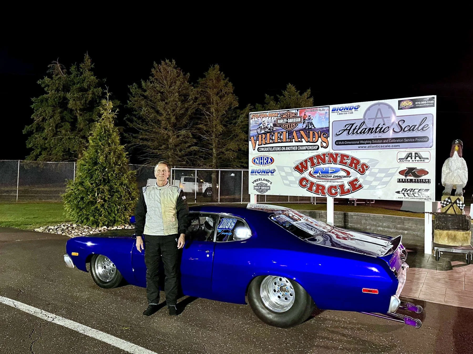 A man in racing gear standing next to a blue drag racing car at night, with a race track and sponsor banners in the background.