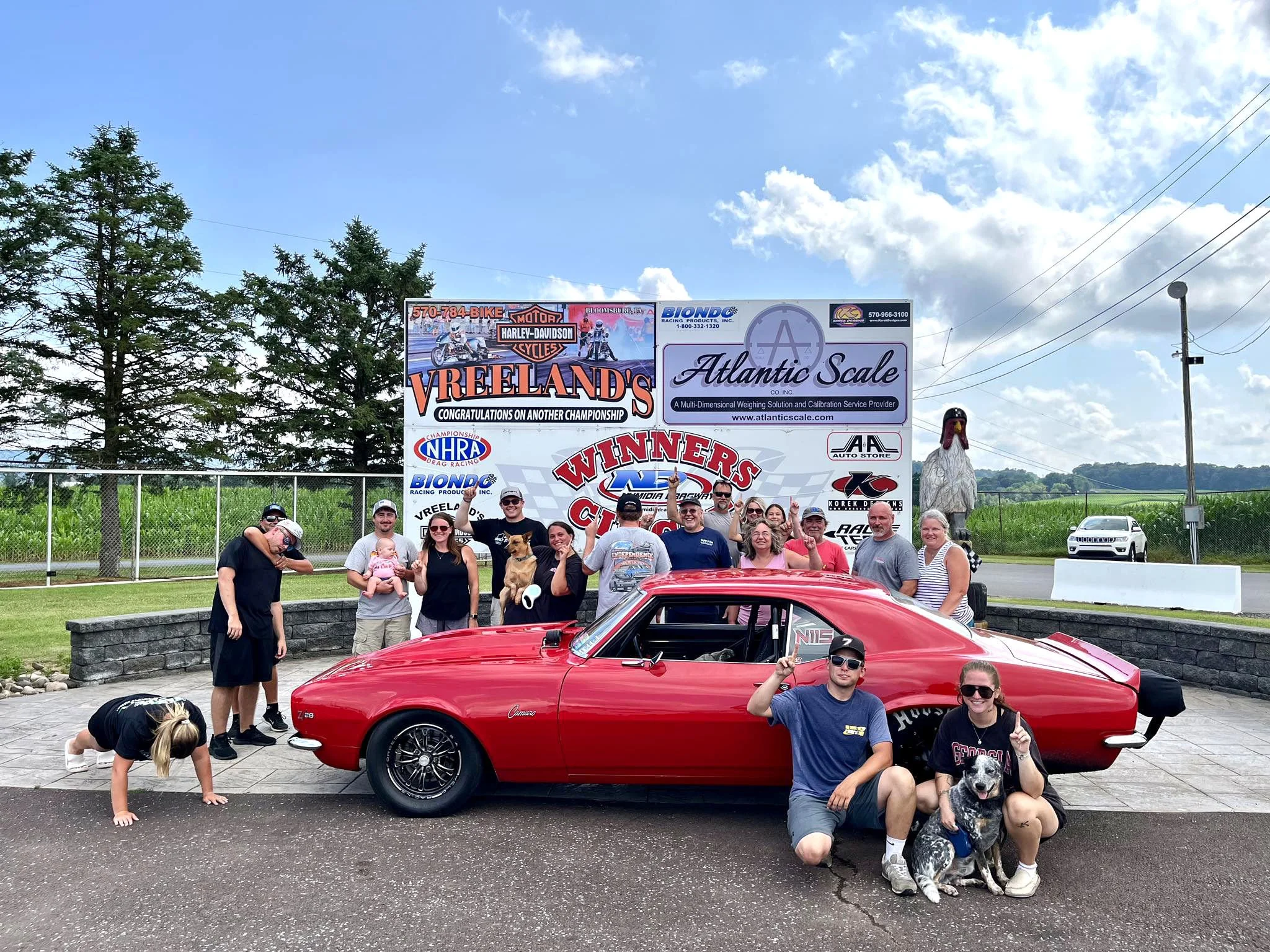 A group of people posing in front of a red classic car at a racing event, with a large signboard and some trees in the background, under a partly cloudy sky.