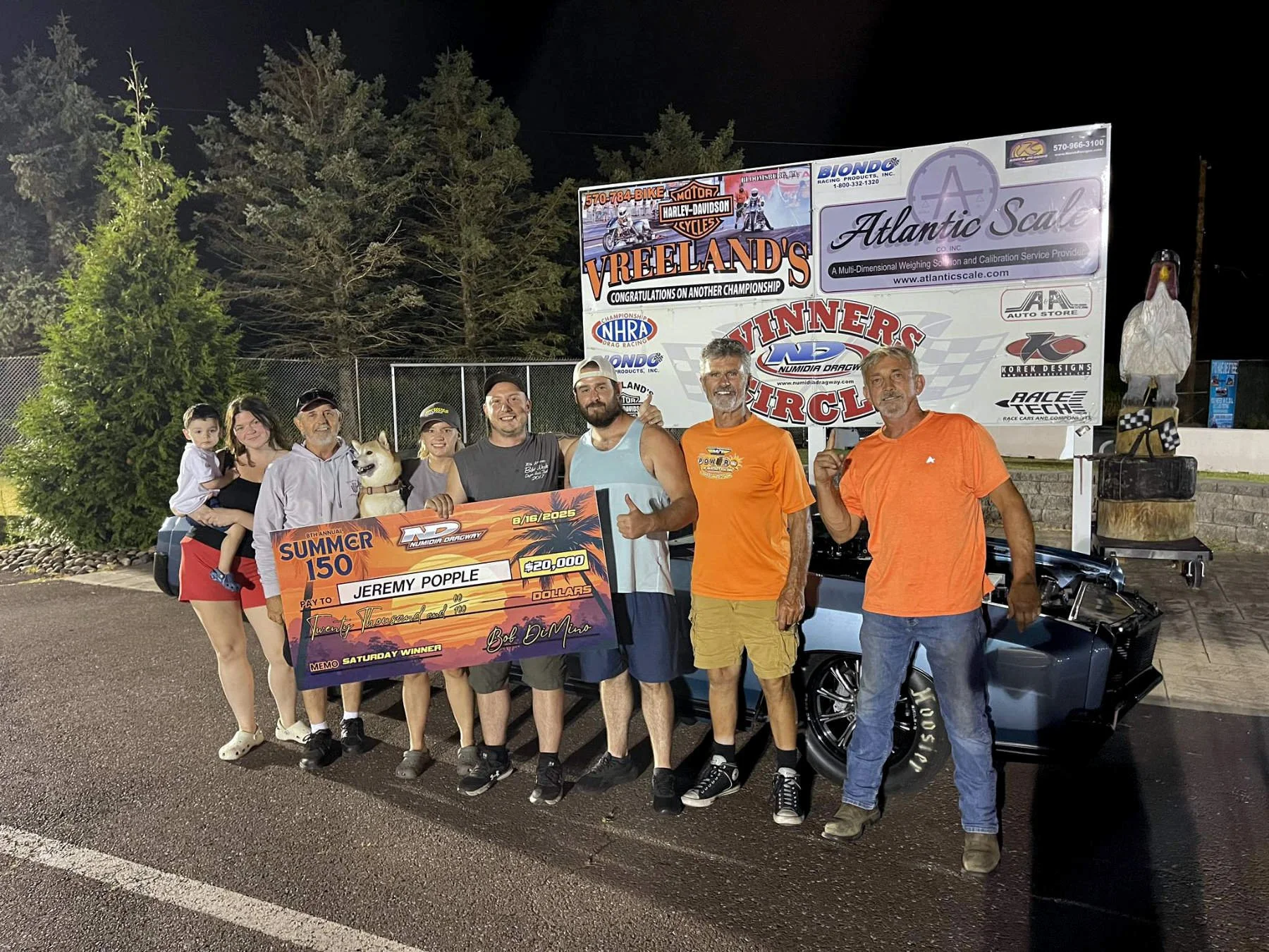 Group of people celebrating at a car racing event at night, holding a large check and a trophy, with a race track sign in the background and a black car nearby.