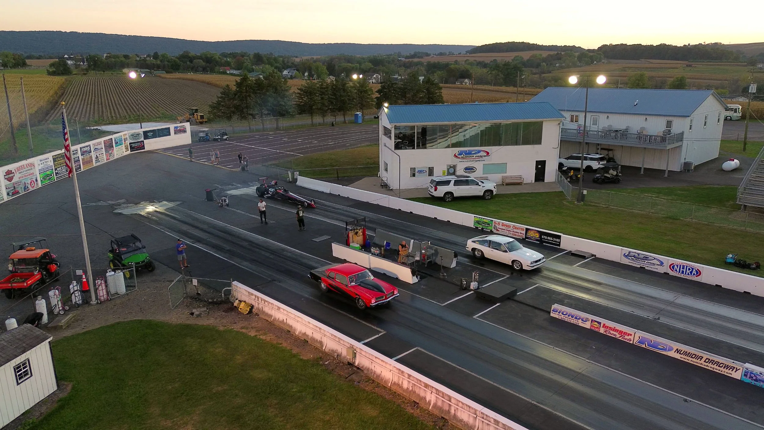 Drag racing track with cars, people, and spectators at sunset. Two cars at the start line, one red and one black, with a small crowd nearby. Large building with signs, parking lot, and scenic countryside in the background.