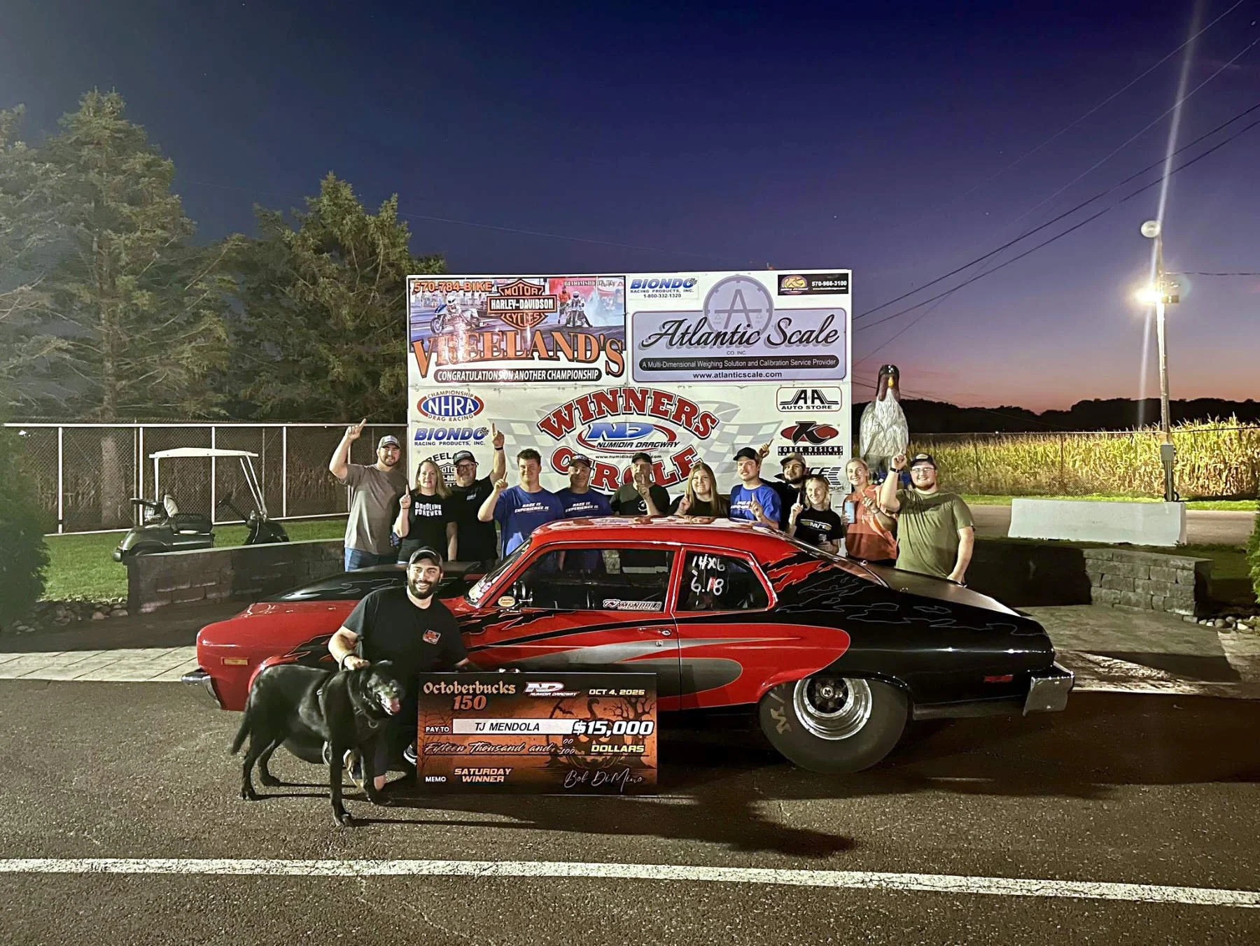 A group of people celebrating a racing victory at night, standing next to a black and red drag racing car with a check for $15,000 in front of them. The backdrop features a large sign with various advertisements, a distant tree line, and an evening s