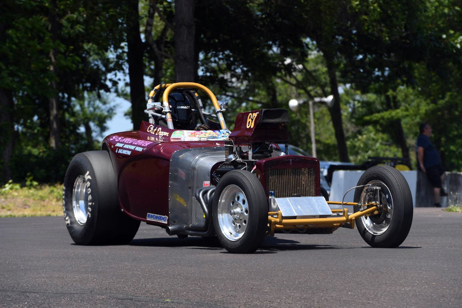A vintage drag racing car with a maroon body, large rear tires, and a roll cage parked on asphalt, with trees and a person standing in the background.