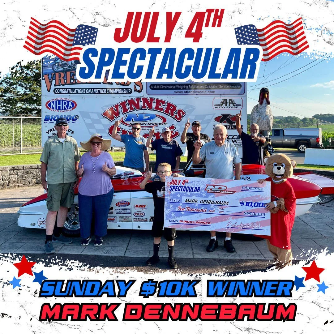 Group of people celebrating open-wheel racing event on July 4th, with a large check for $10,000 awarded to Mark Dennebaum, front and center holding the check, standing next to a racing car and mascot, with a banner and American flags in the backgroun