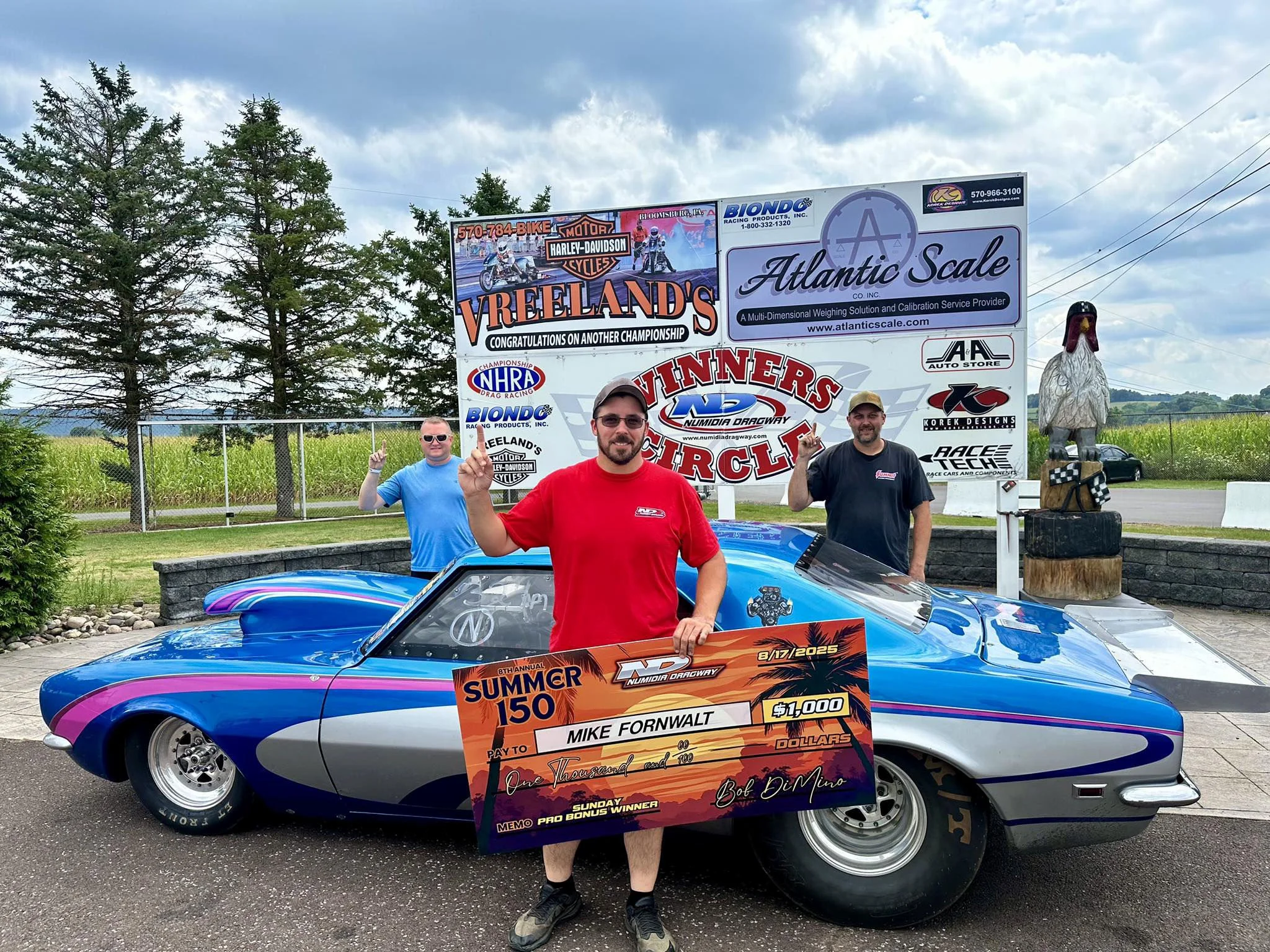 Three men standing next to a blue and gray race car, holding a large orange check for $1,000 for winning a summer race at Numidia Dragway, with a sign and trees in the background.
