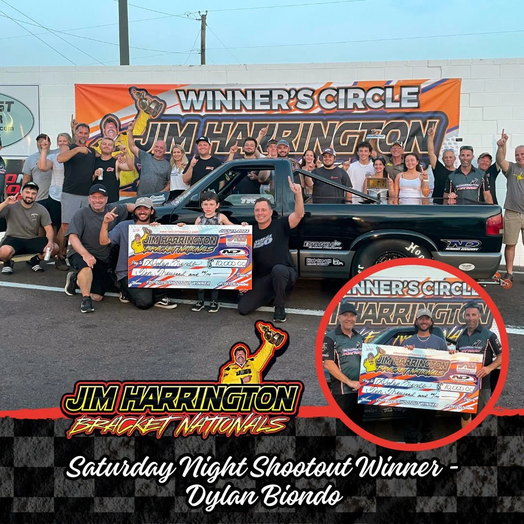 Group of people celebrating a racing victory, holding a large check and raising their hands, with a black racing pickup truck and a race track banner in the background.