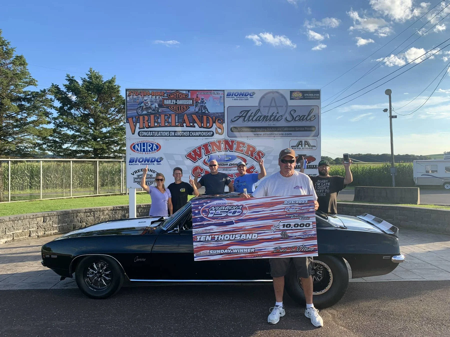 Group of people standing beside a black muscle car with a large celebratory check and racing banners in the background. The scene appears to be at a racing event or celebration.