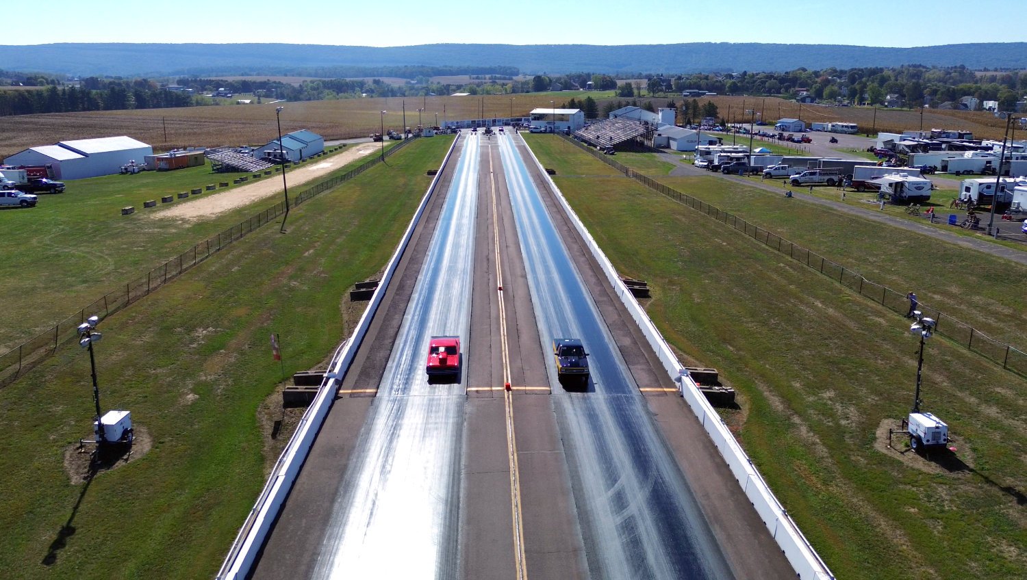 Two race cars on a drag strip during a race in a rural area with several tents, vehicles, and spectators along the sides of the track.