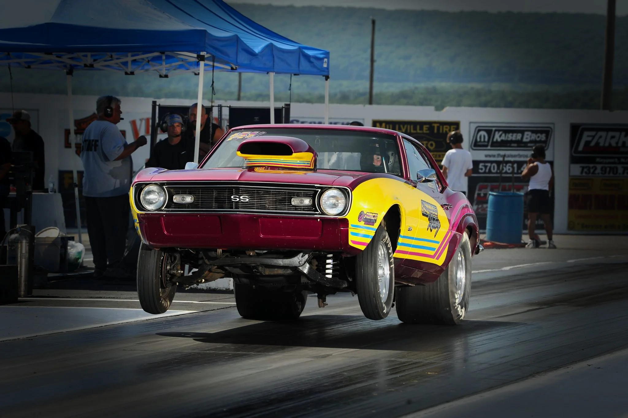 A racing car with a bright pink and yellow paint job is lifted off the ground during a drag race at a racetrack. The car has large rear tires and a prominent hood scoop. Several crew members and officials are present in the background under a blue ca