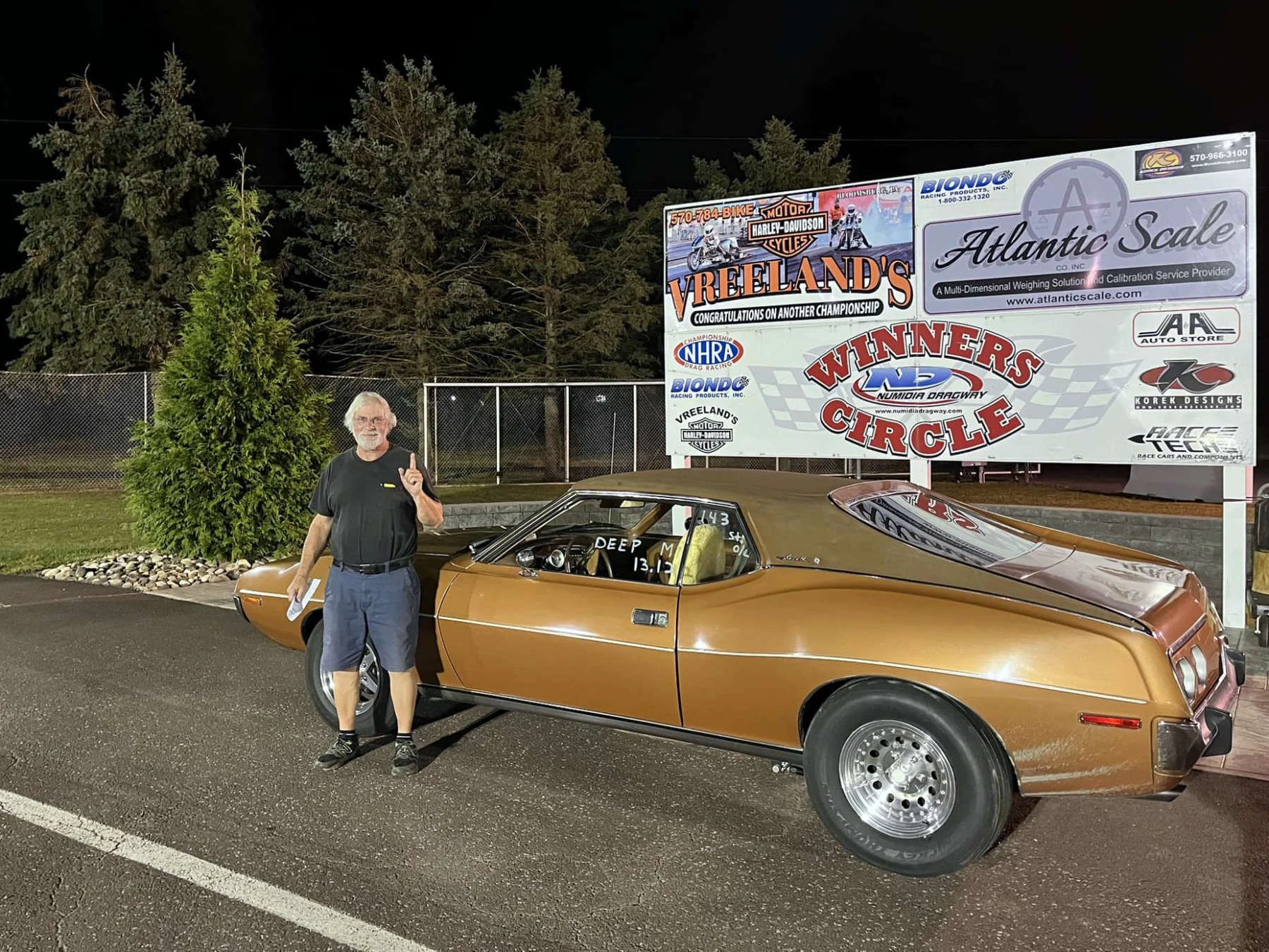 A man with gray hair, a beard, wearing a black t-shirt, blue shorts, and black shoes, standing next to a vintage brown muscle car at night. Behind him is a tall green tree, a chain-link fence, and a large billboard with various advertisements, includ