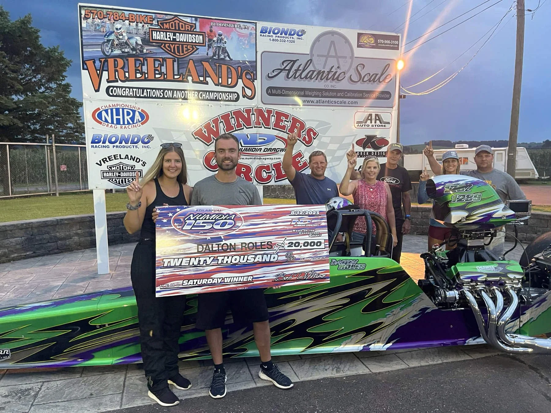 A group of six people celebrating a drag racing victory at the Nemuda Dragway with a large check for twenty thousand dollars awarded to Dalton Roles, standing behind a green and purple race car with a building and signboards in the background.