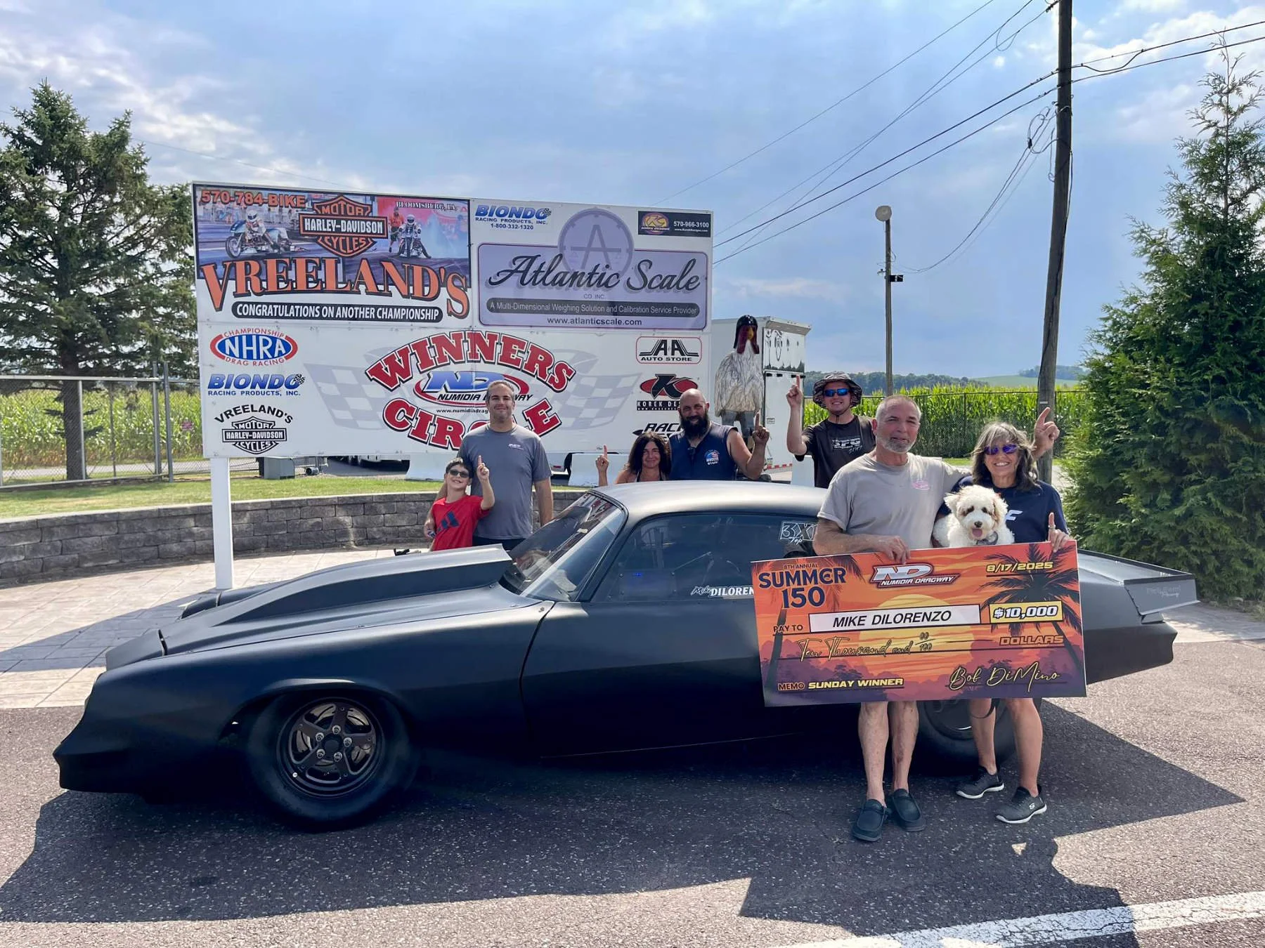 A group of seven people, including children and adults, posing with a black race car on an outdoor street during daytime. They are celebrating a victory, holding a large check for $10,000 and a trophy. Behind them are commercial signs including Vreel