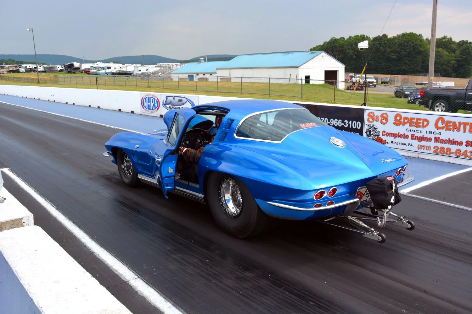 A vintage blue race car on a drag strip with driver preparing for a race. The car has a rounded, aerodynamic body with four taillights and is positioned at the starting line. The background includes track advertisements, fencing, a large building, an