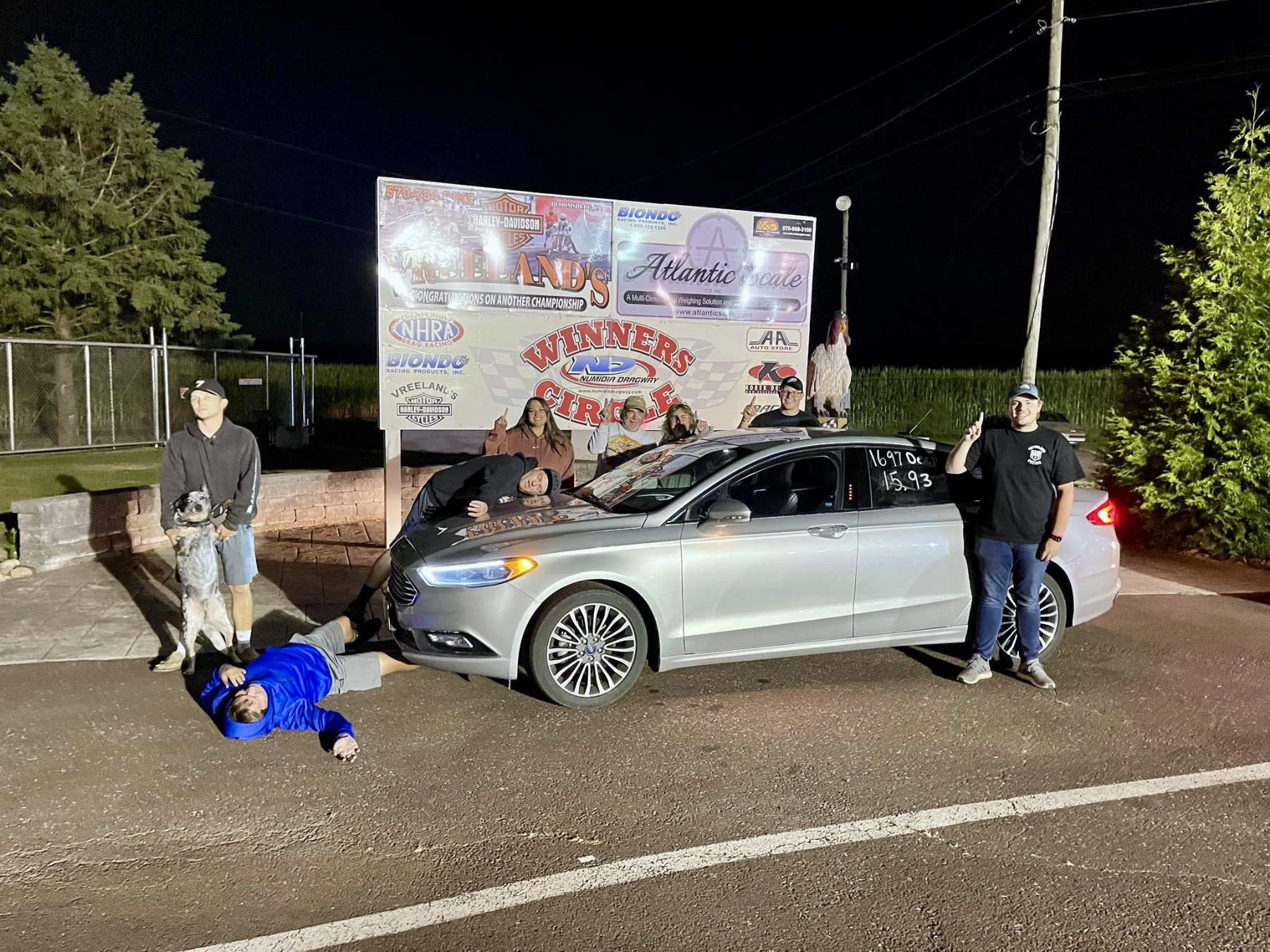 Nighttime scene with six people and a dog around a silver car in front of a large sign. One person lies on the ground in front of the car, another is leaning on it, three are standing, and one person is holding up a finger. The sign behind them has v