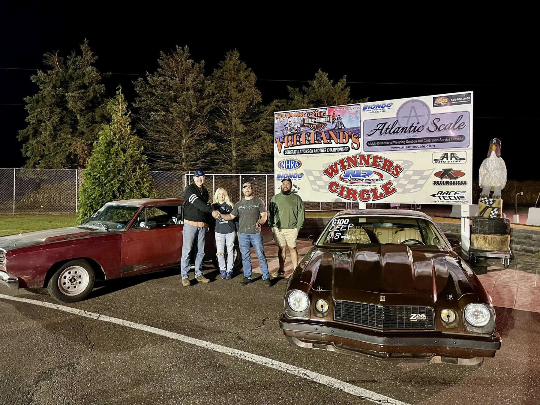 Four people standing together in a parking lot at night, posing with two vintage cars. Behind them, a large sign reads 'Winners' Circle' and features various logos and advertisements, with several trees in the background.