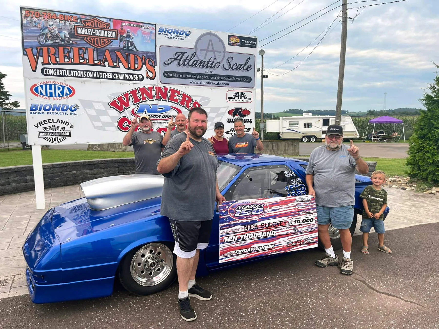 Group of five people, including a young boy, celebrating next to a blue race car with a large check for ten thousand dollars, in front of a Harley-Davidson dealership sign, with the man in front holding up one finger.