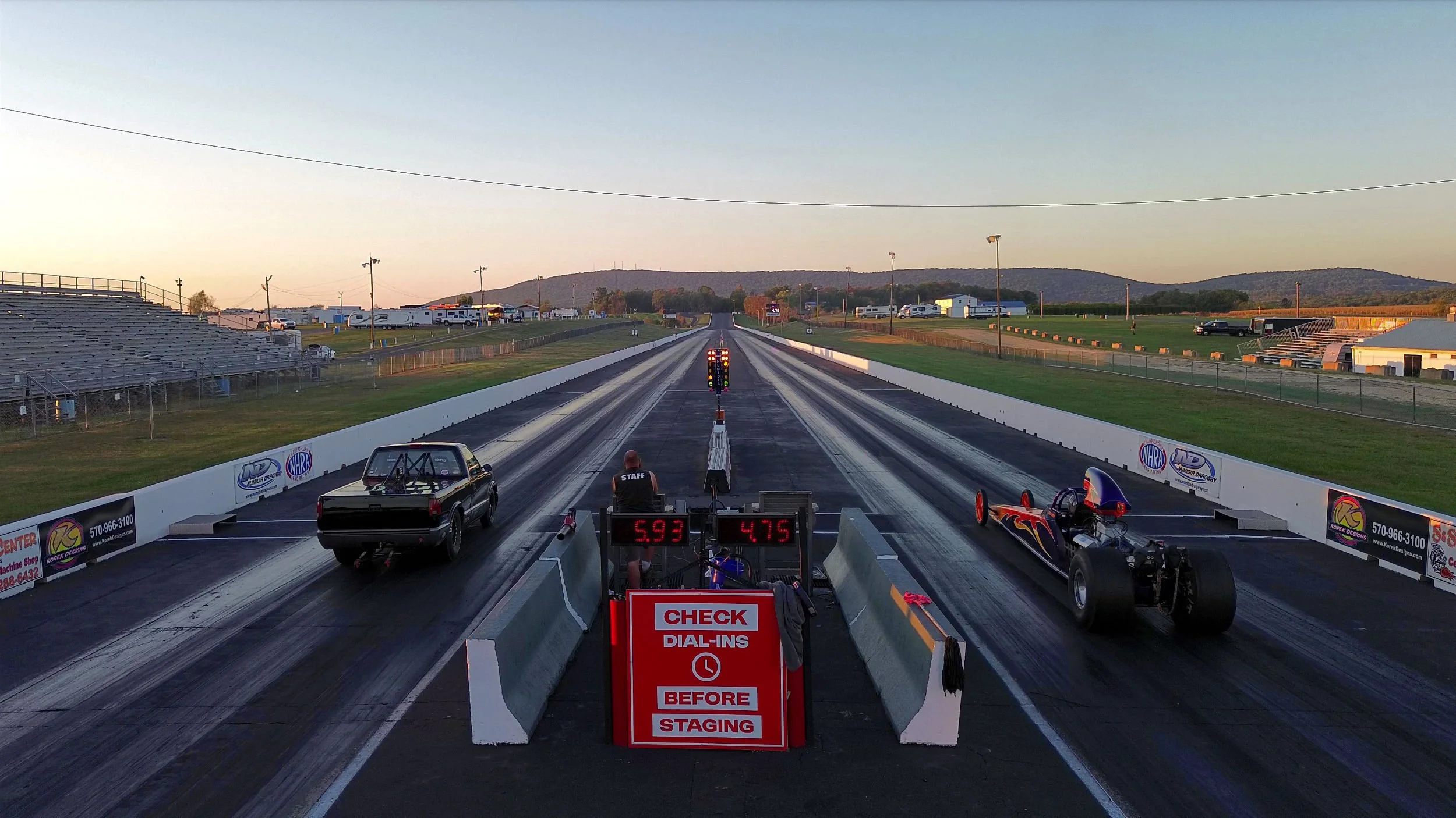 Drag racing track at sunset with cars, race officials, and staging area.