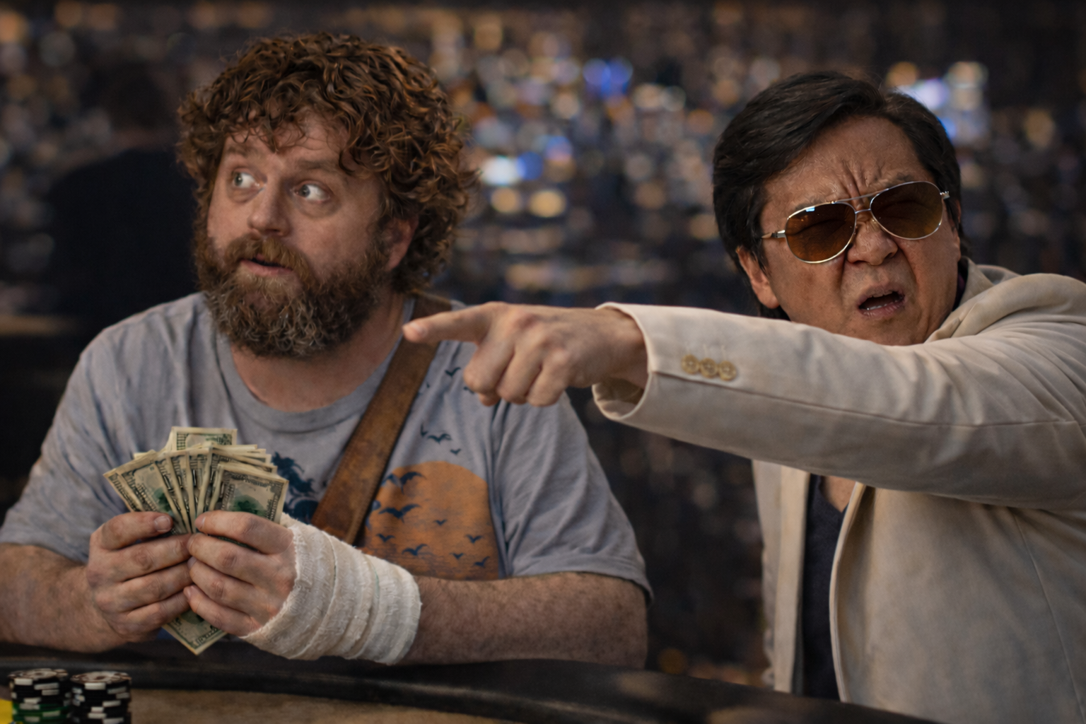 Two men at a poker table, one with a stack of cash and a bandaged hand, looking confused, and the other wearing sunglasses, pointing and speaking.