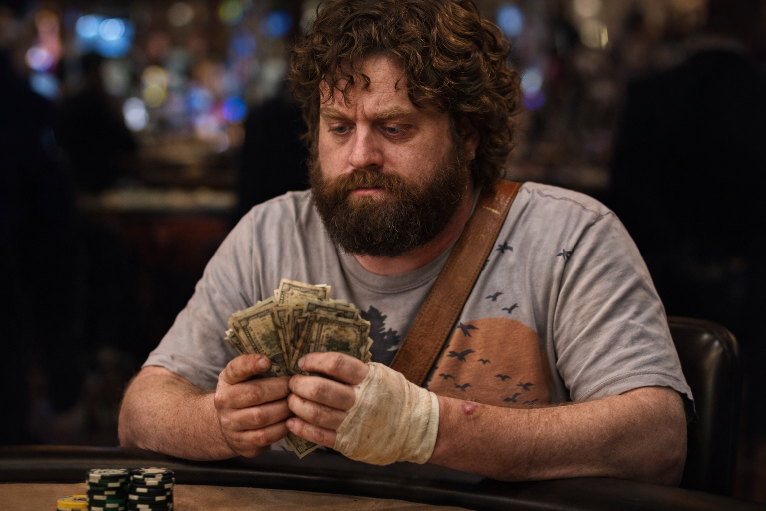 A man with curly hair and a beard, wearing a gray t-shirt with a sunset and bird design, is sitting at a poker table counting a large stack of dollar bills. There are poker chips in front of him, and the background is blurred with people in a dimly lit setting.