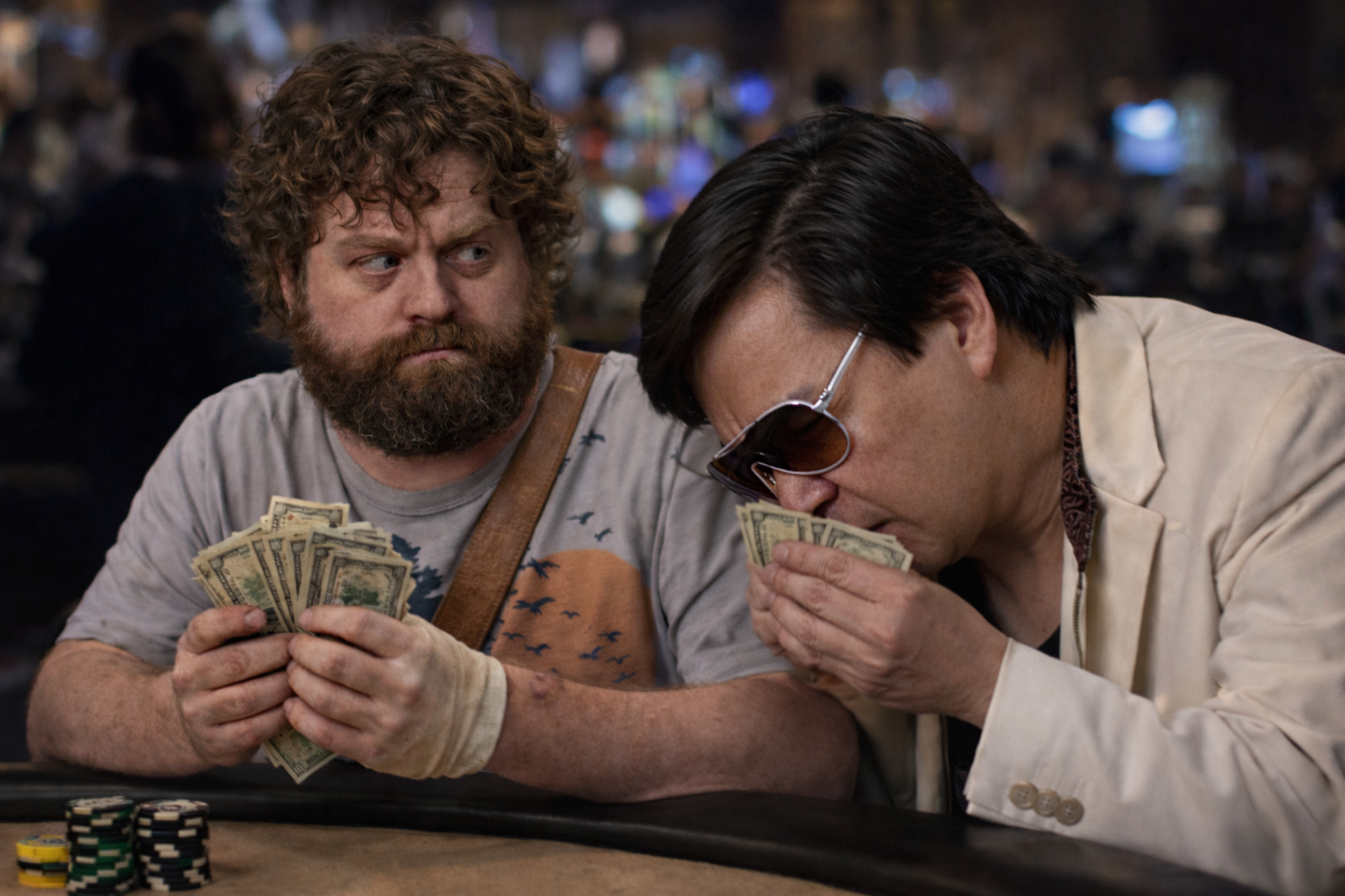 Two men sitting at a poker table, one with curly hair and beard holding cash, the other wearing sunglasses and a cream-colored blazer kissing cash, with poker chips stacked on the table.