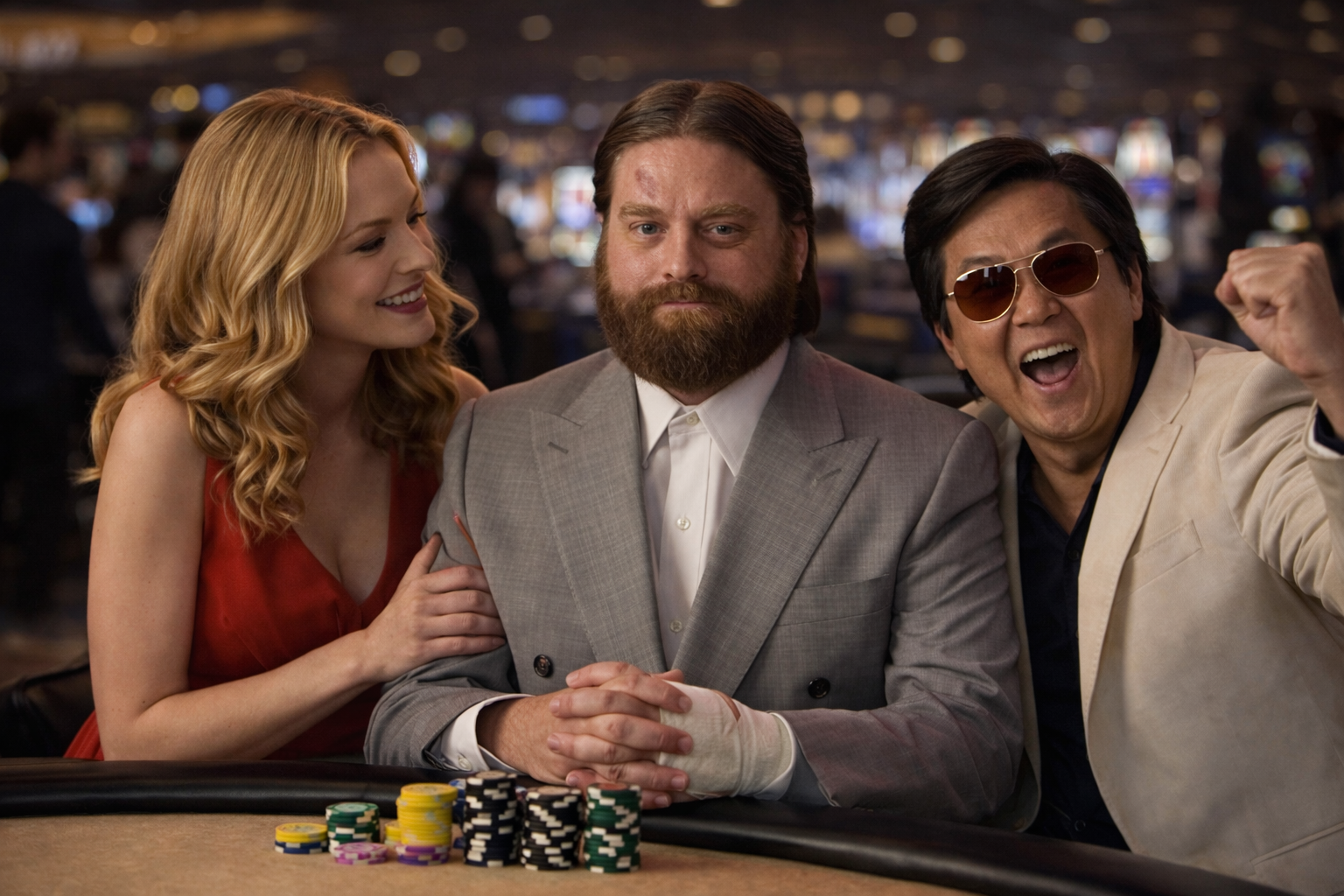 Three people at a casino poker table, two men and a woman. The woman has long curly blonde hair and is wearing a red dress, smiling and touching the man in the middle. The man in the middle has a beard, is dressed in a gray suit, and appears serious. The man on the right has dark hair, wearing sunglasses and a cream-colored jacket, cheering with a raised fist.