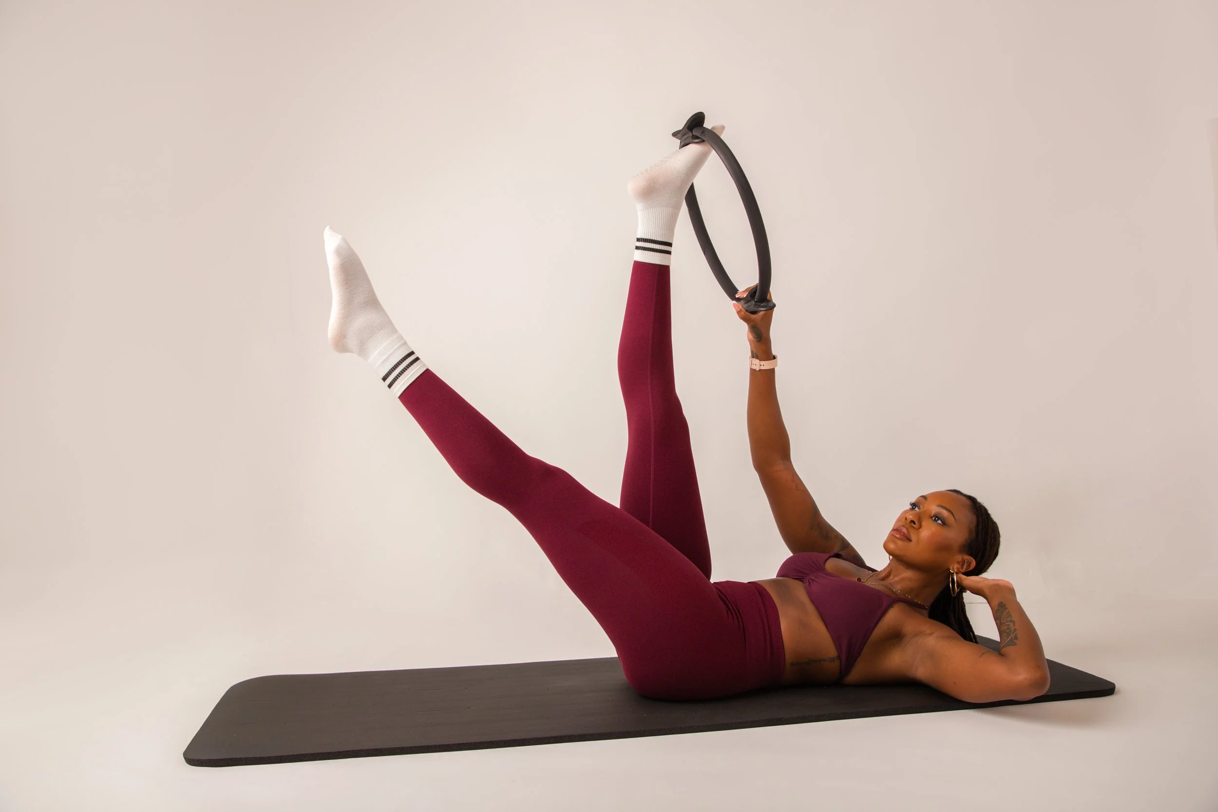 A woman in sportswear lying on a black yoga mat, holding a fitness ring with one arm, raising her leg, and resting her head on her other arm, against a plain white background.