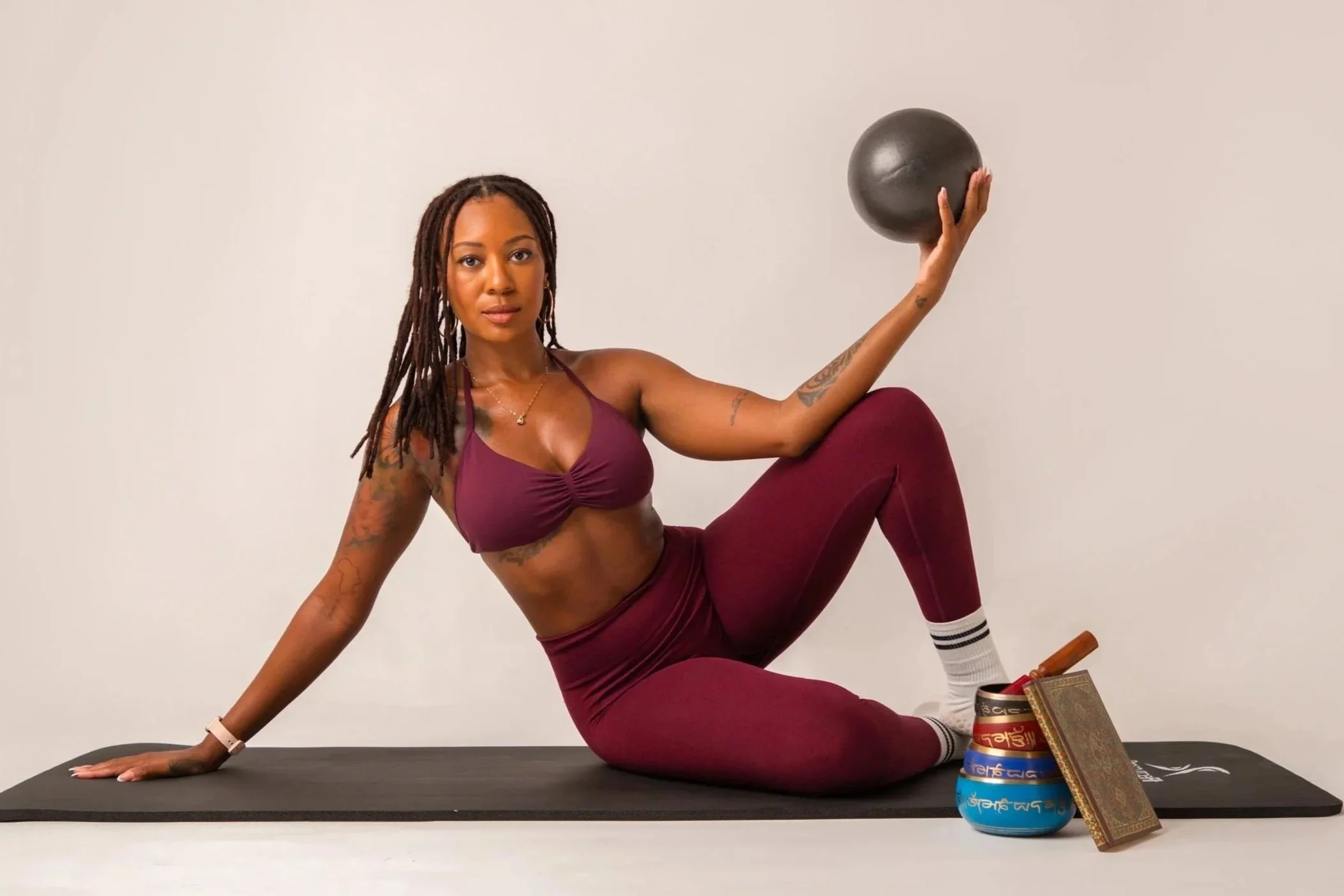 A woman with dreadlocks exercising on a black yoga mat, holding a black medicine ball in her raised right hand, wearing a burgundy sports bra and matching leggings, with yoga props including a stack of singing bowls and a playing stick next to her.