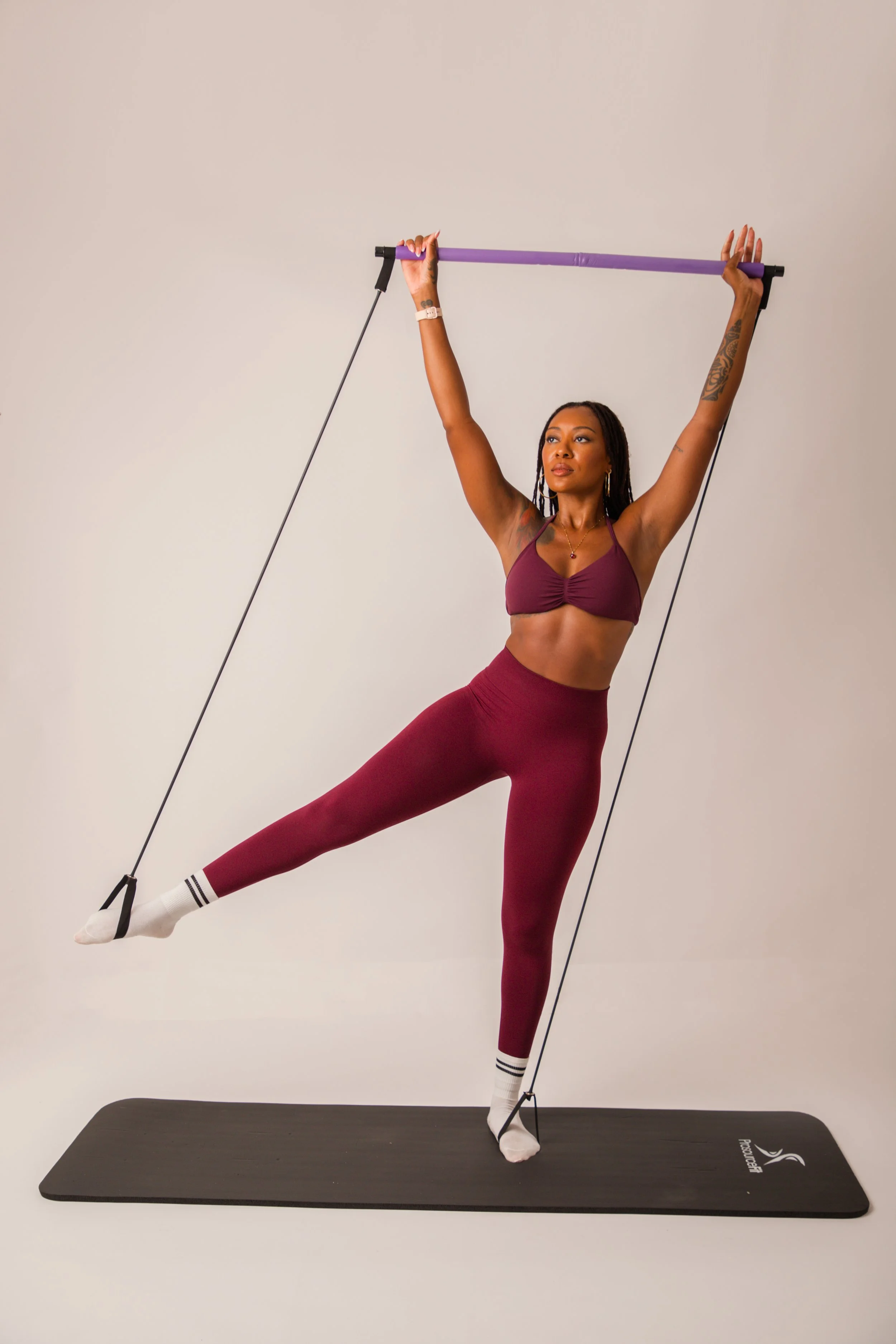 Woman in burgundy sportswear performing a balance yoga pose with resistance band, standing on a black yoga mat in front of a plain white background.