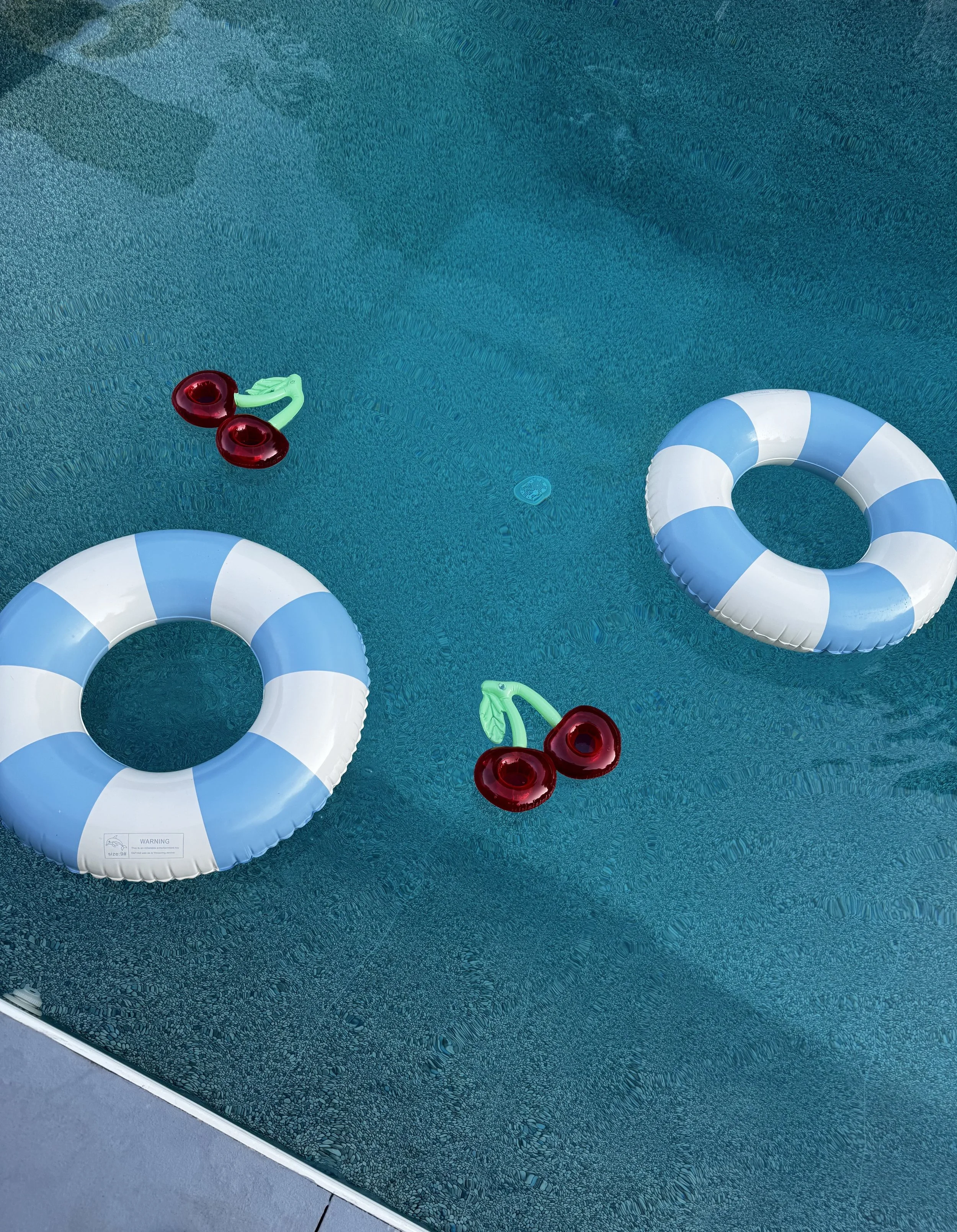 Two blue and white striped pool float rings and two red cherry-shaped pool toys float on a swimming pool with clear water.