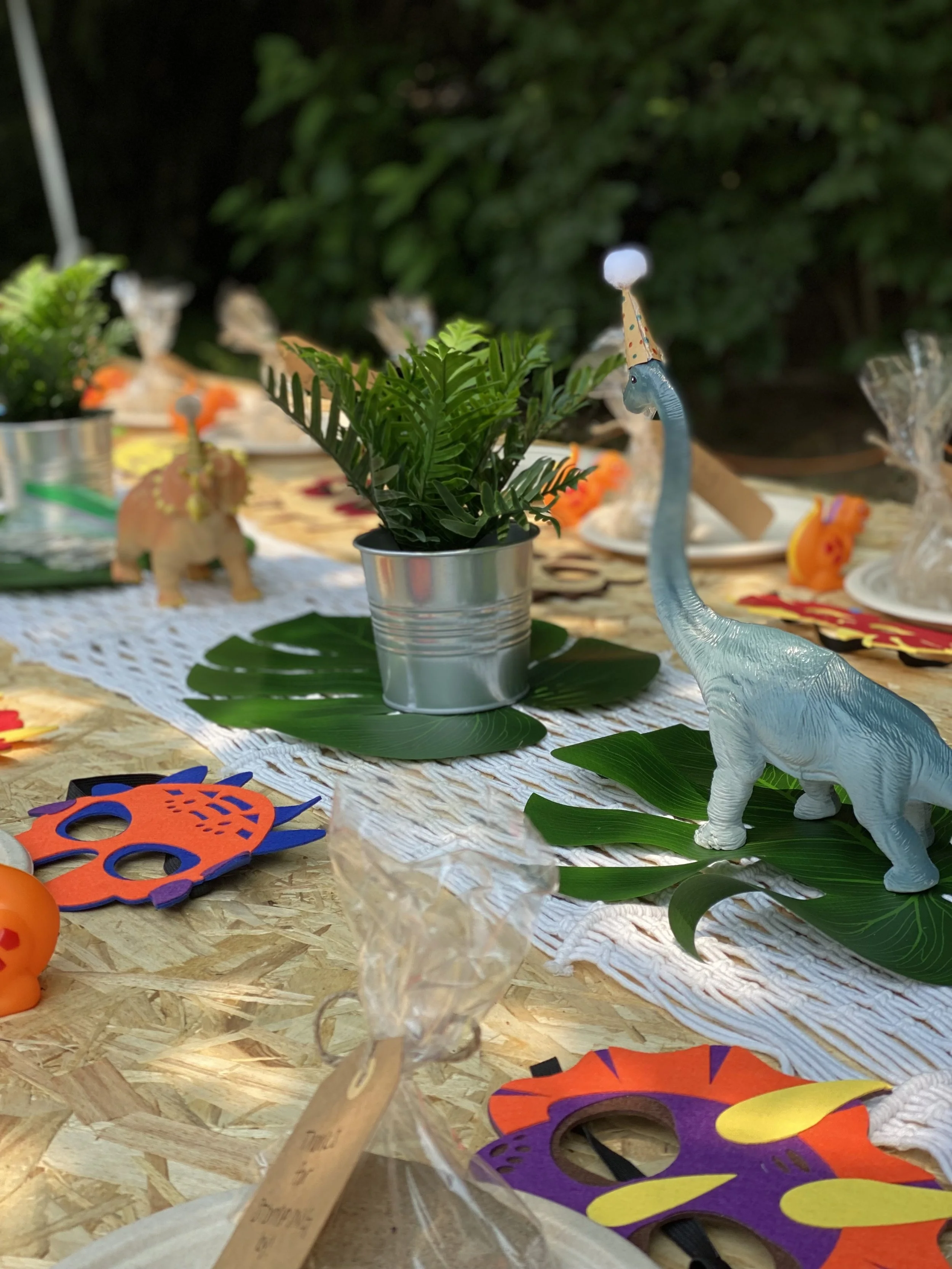 Decorated outdoor table with tropical and dinosaur-themed party decorations, including a small potted plant, a plastic dinosaur wearing a party hat, paper masks, and wrapped candies, with a lush green background.