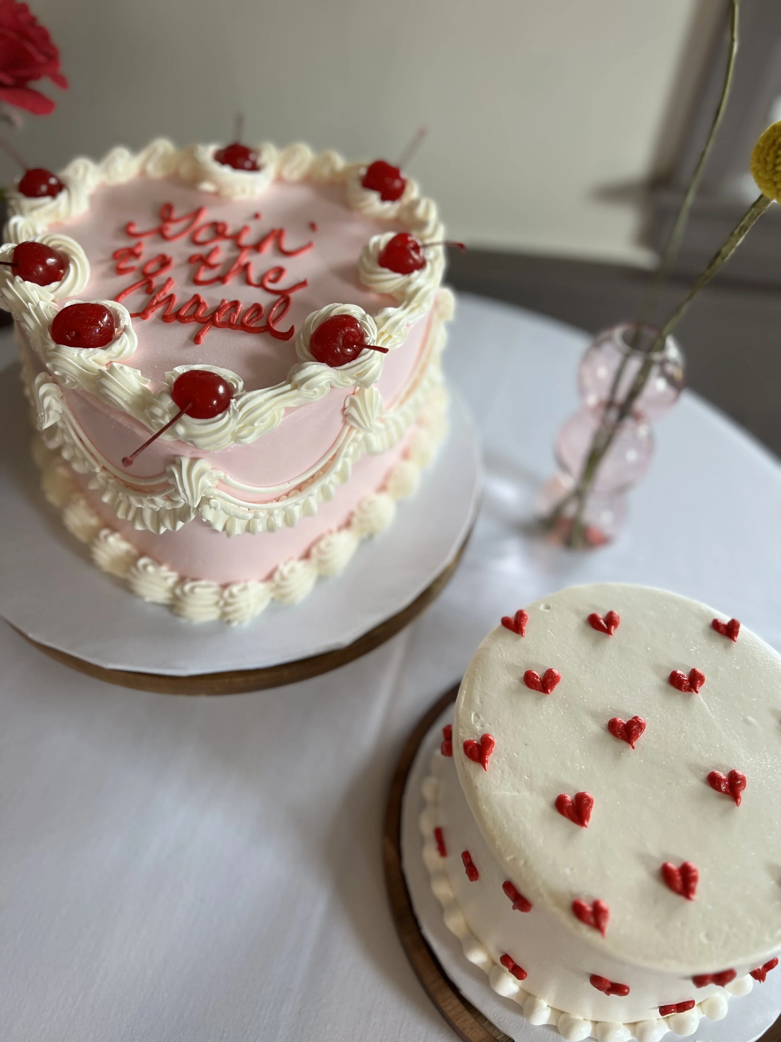 Pink heart-shaped cake with white icing, cherries on top, and red writing that says 'Main on the chapel.' Next to it is a round white cake decorated with red hearts, placed on a party table with a white tablecloth and simple vases with yellow and pin