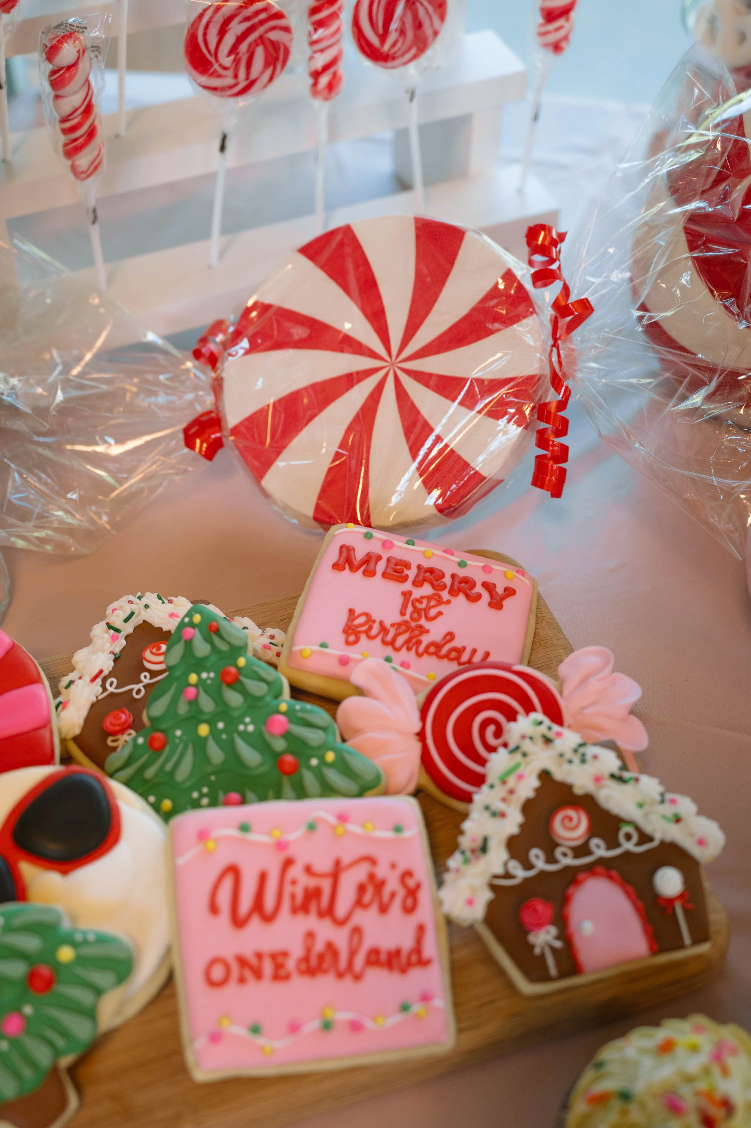 Decorated Christmas cookies on a wooden tray, including a gingerbread house, a Christmas tree, and cookies with birthday messages, with Christmas candies in the background.