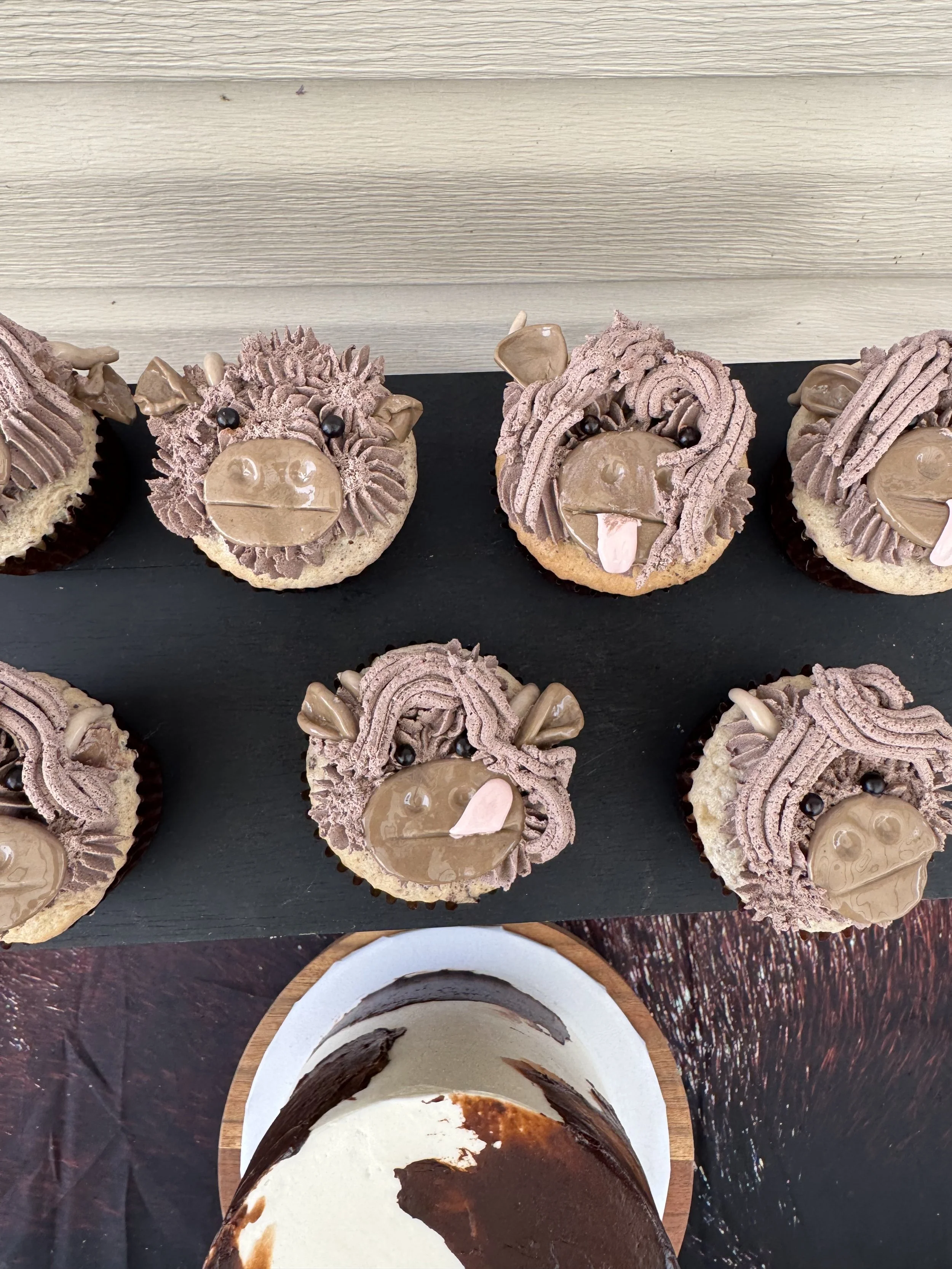 Six cupcakes decorated to look like hippos, with chocolate frosting, candies, and icing details, displayed on a black board. A partially eaten cake is visible at the bottom.