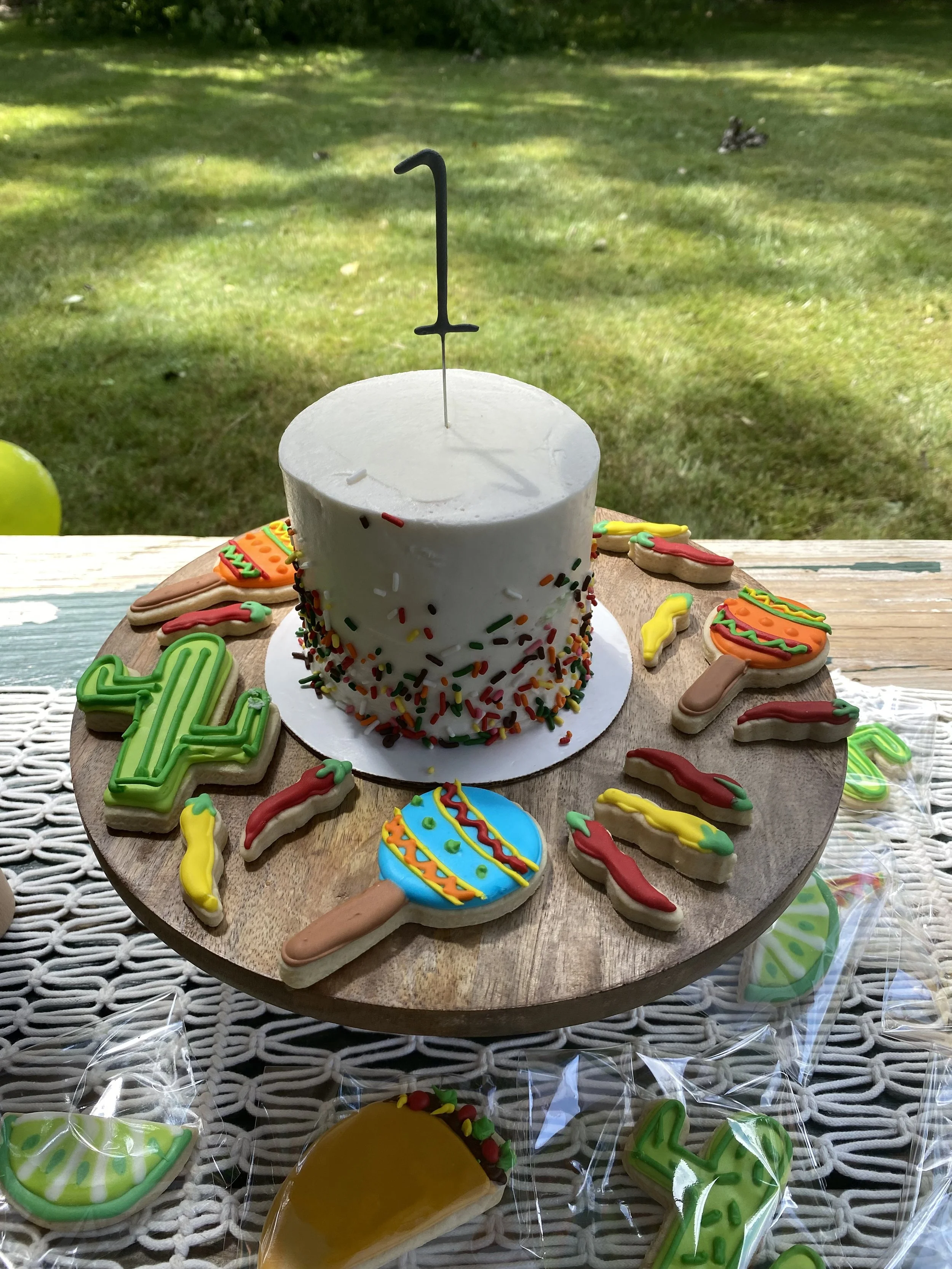 A white birthday cake with colorful sprinkles on a wooden cake stand, decorated with a black number '1' candle. Surrounding the cake are festive cookies shaped and decorated like tacos, chips, and lollipops, with some wrapped in plastic. The scene is