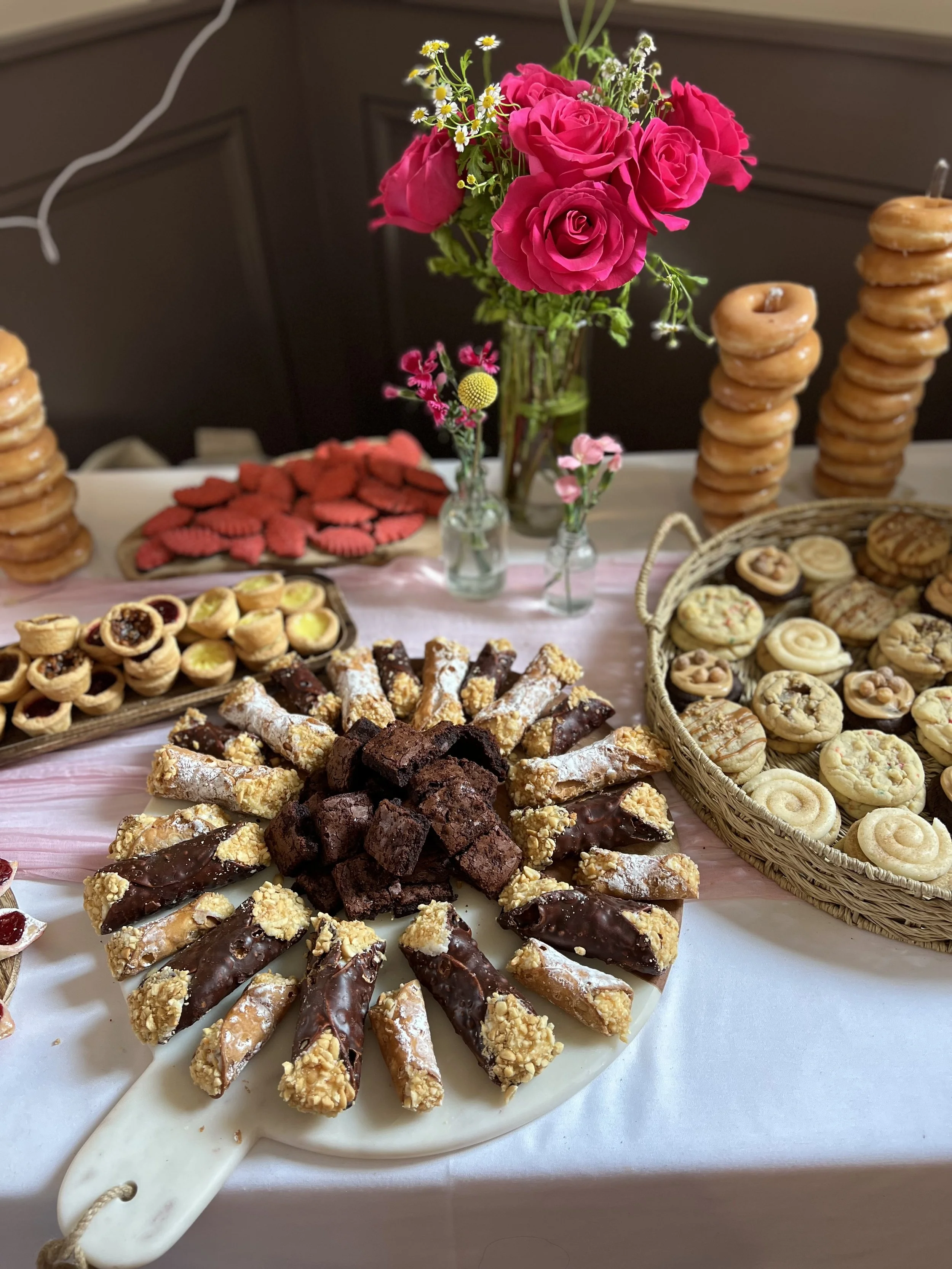 Sweet treats and baked goods on a table with pink roses in glass vases in the background.
