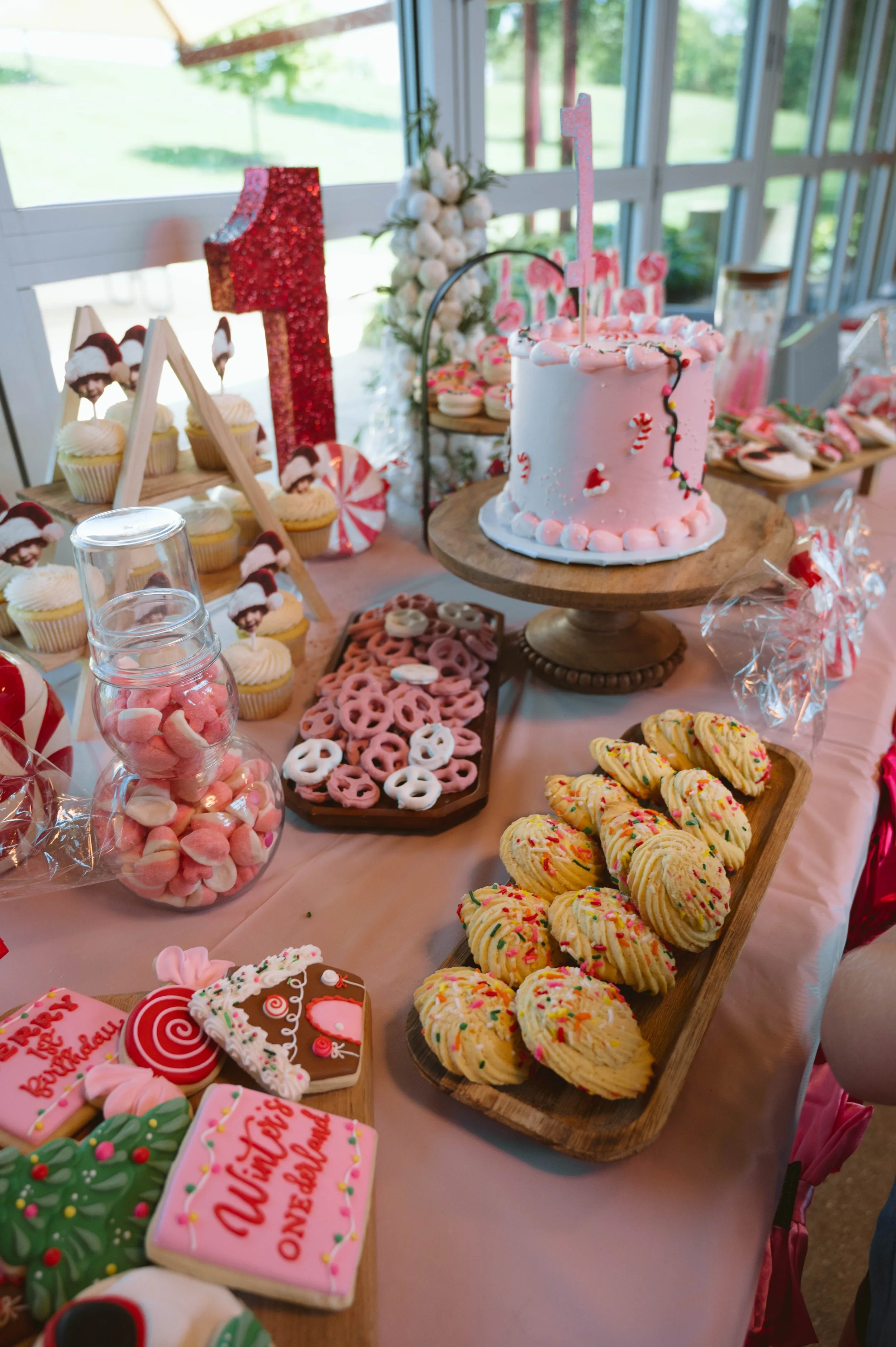 Party table with pink and white themed cookies, cupcakes with Santa hats, a pink birthday cake with a number 1 candle, and assorted candies, celebrating a first birthday.